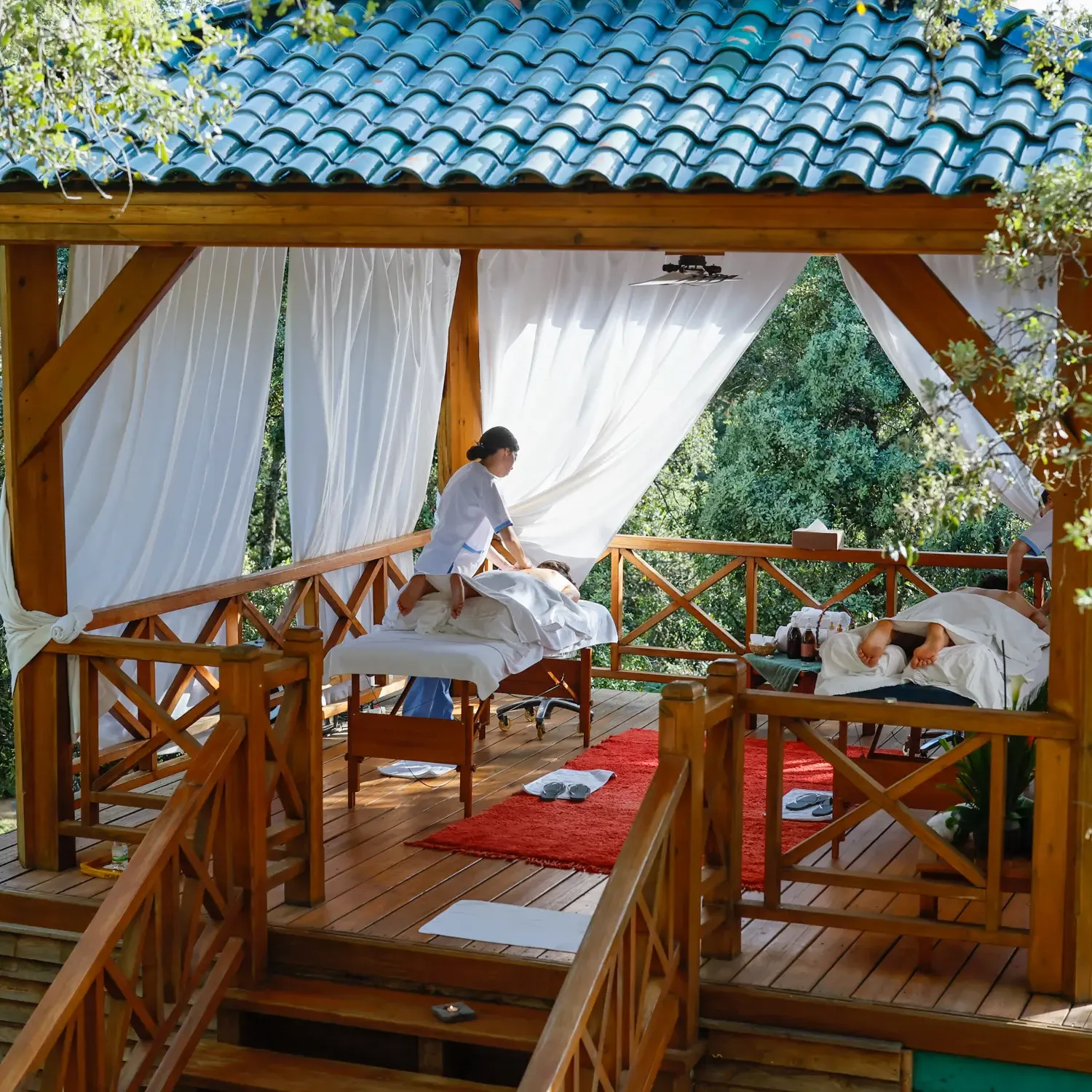 Deux personnes allongées sur des tables de massage en plein air sous un auvent en bois avec un toit en tuiles bleues, entourées d'arbres