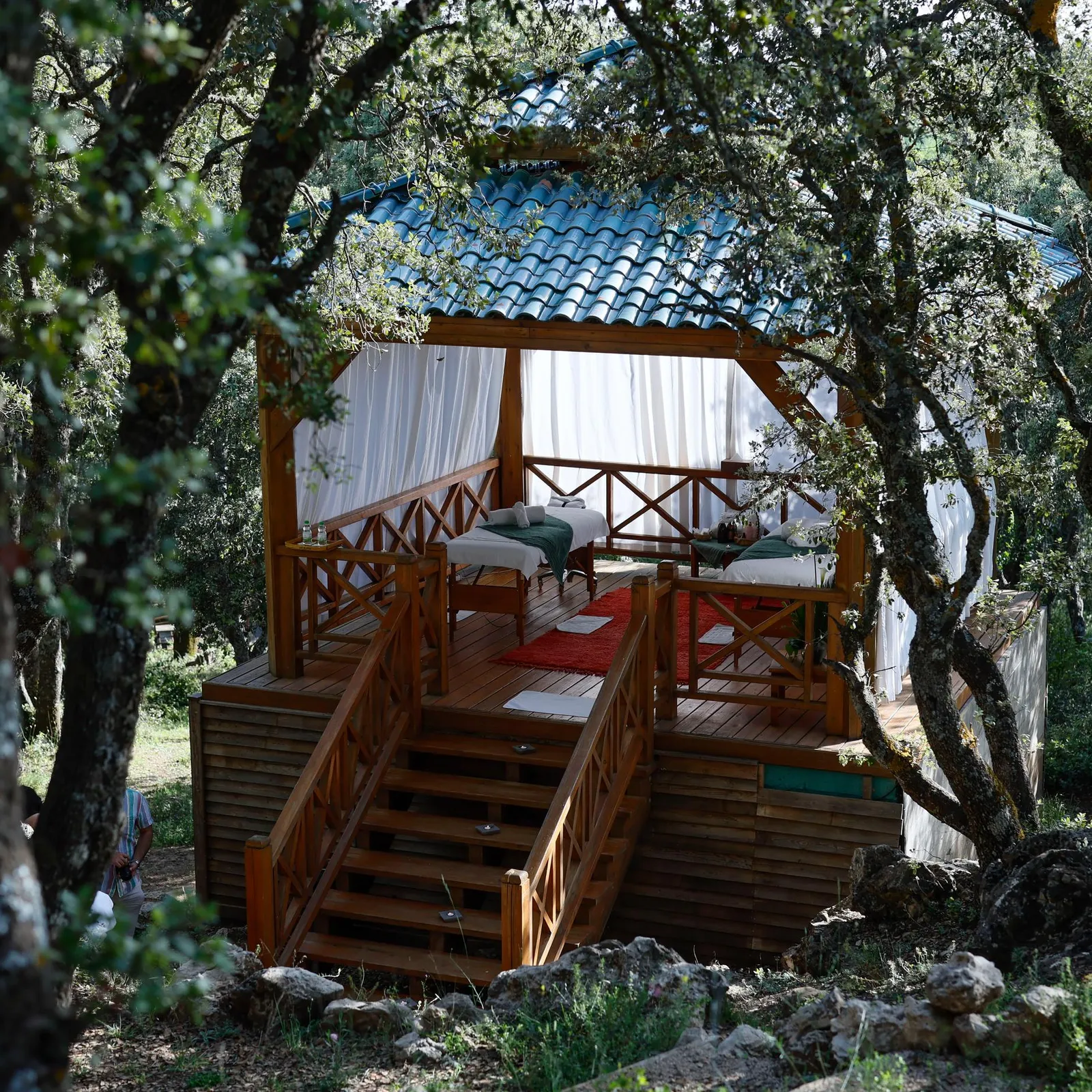 Une cabane en bois avec un lit et des coussins est située dans une forêt, entourée d'arbres et accessible par un escalier