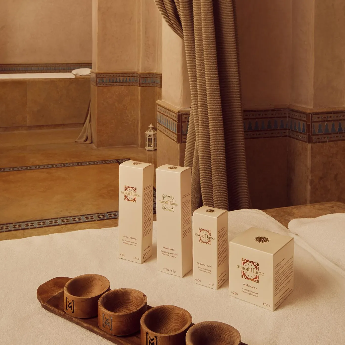 Quatre boîtes de produits cosmétiques et un plateau en bois avec des coupelles posés sur un tapis blanc dans une pièce aux murs ornés de carreaux de céramique