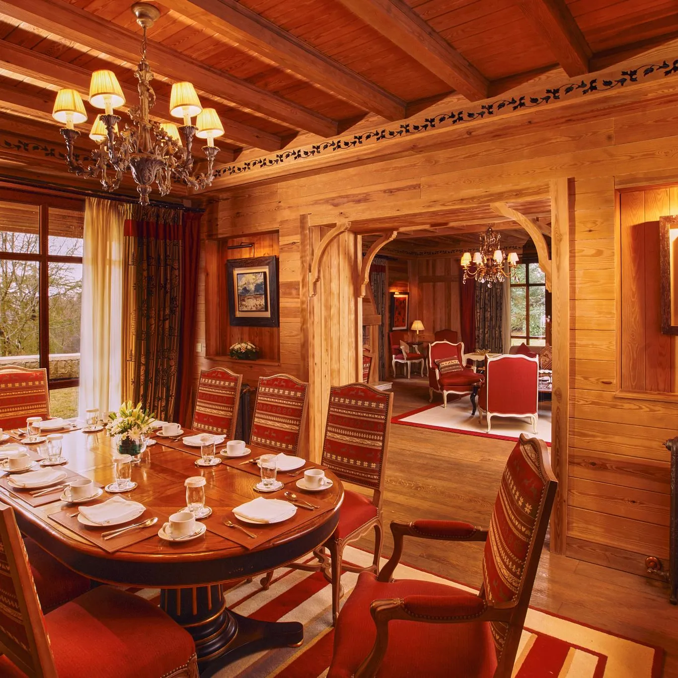 Salle à manger en bois avec une grande table ronde, des chaises rouges et des chandeliers, donnant sur un jardin à travers de grandes fenêtres