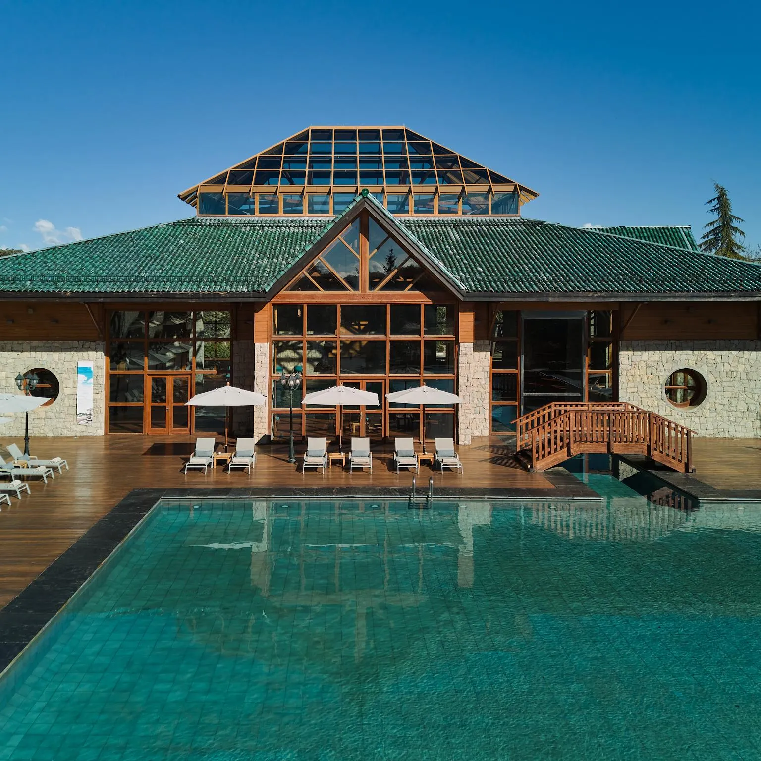 Un bâtiment en pierre et bois avec un toit vert et une extension vitrée, devant une piscine extérieure entourée de chaises longues et de parasols