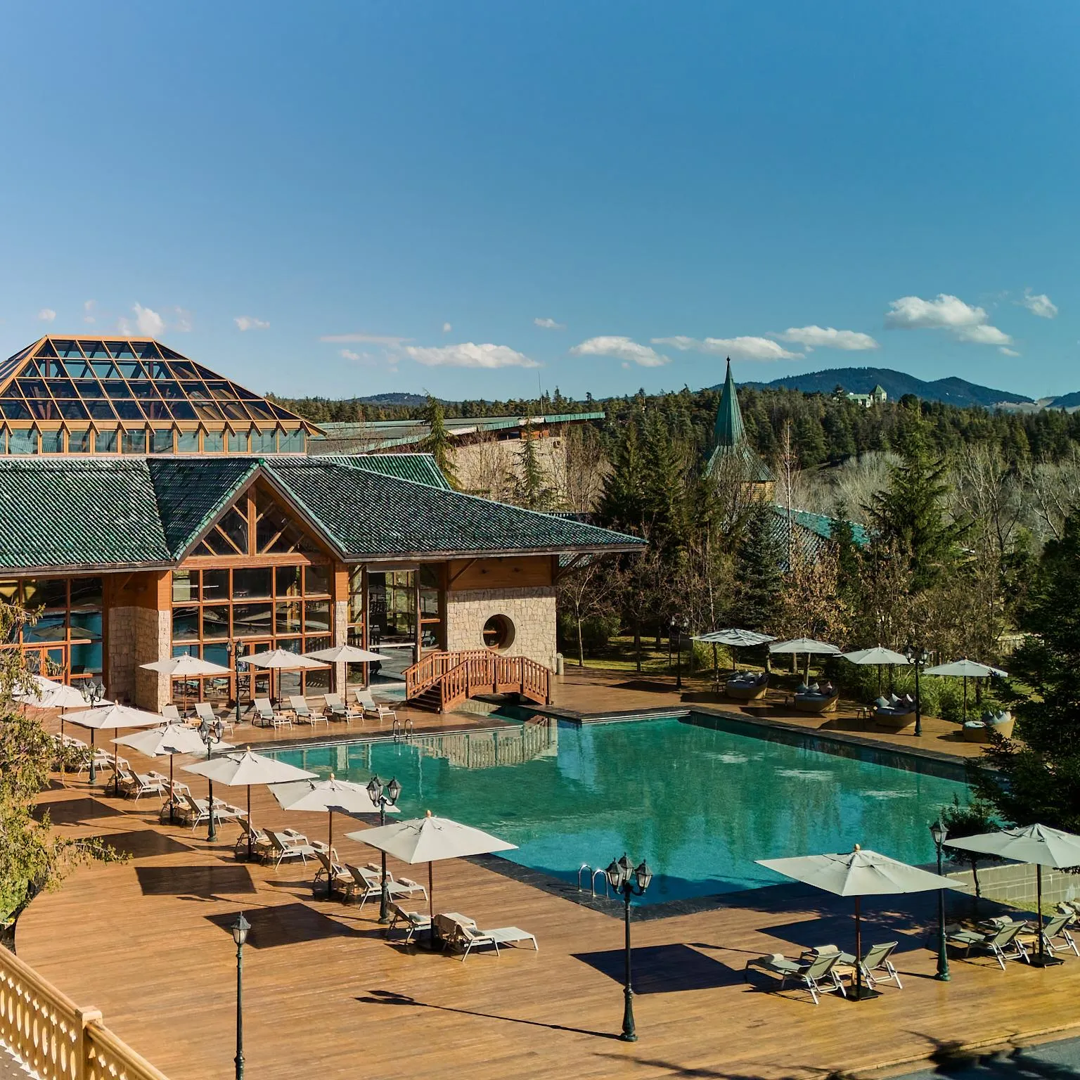 Piscine extérieure entourée de transats et de parasols, avec un bâtiment en bois et toit vert en arrière-plan, dans un cadre naturel montagneux