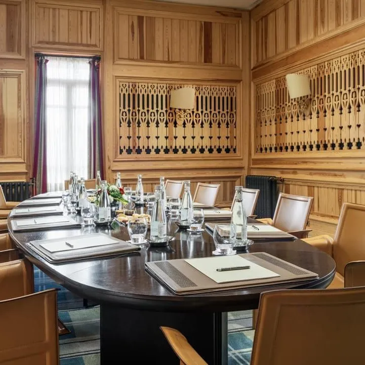 Salle de réunion avec une table ovale entourée de chaises en cuir, des verres et des carnets posés sur la table, des murs en bois avec des motifs sculptés et des fenêtres avec des rideaux