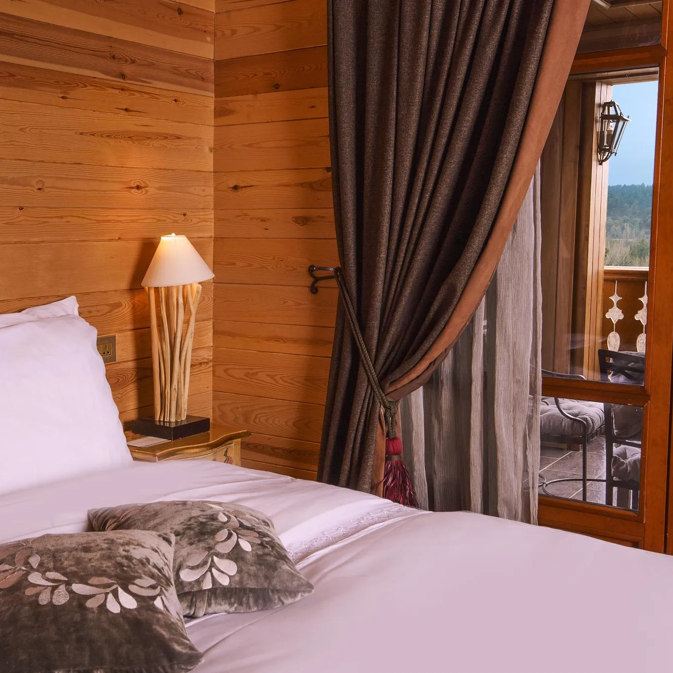 Chambre en bois avec un lit, des coussins, une lampe de chevet et une porte-fenêtre ouverte sur un balcon avec vue sur un paysage