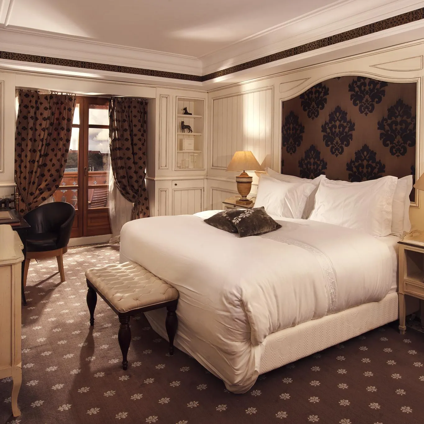 Chambre élégante avec un grand lit blanc, des tables de chevet, un bureau, une chaise et une porte-fenêtre donnant sur un balcon