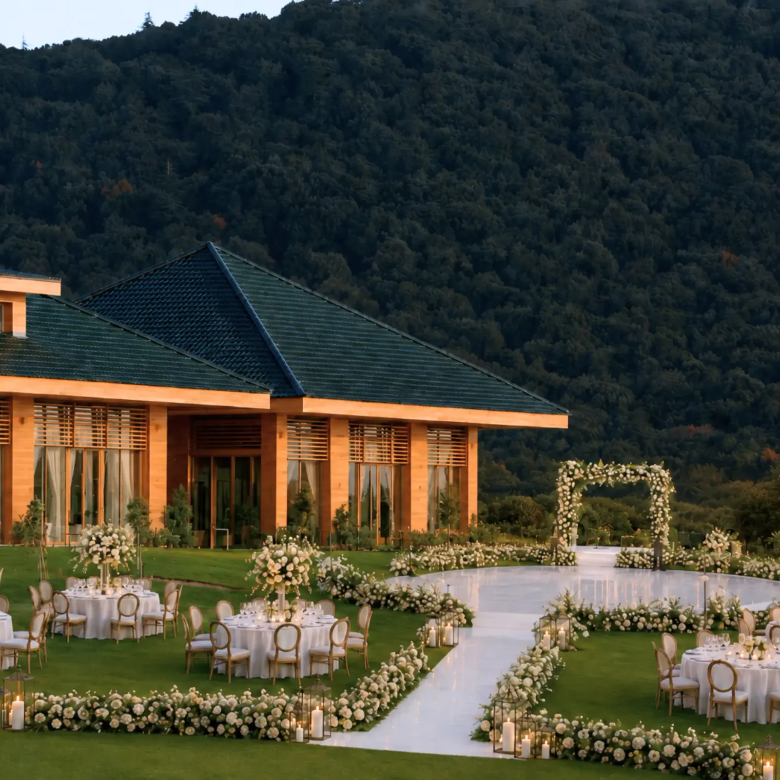 Un espace extérieur aménagé pour un événement avec des tables, des chaises et des décorations florales, devant un bâtiment aux murs orange et toit vert, entouré d'une colline boisée