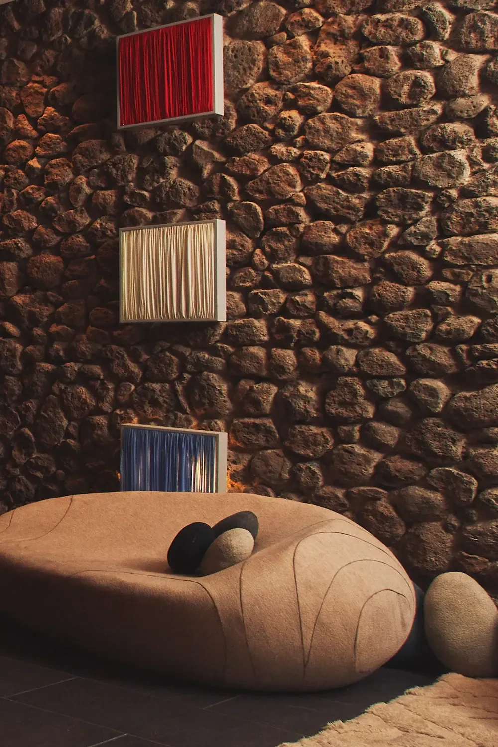 A shell-shaped sofa with cushions sits in front of a stone wall with three paintings in red, beige, and blue.