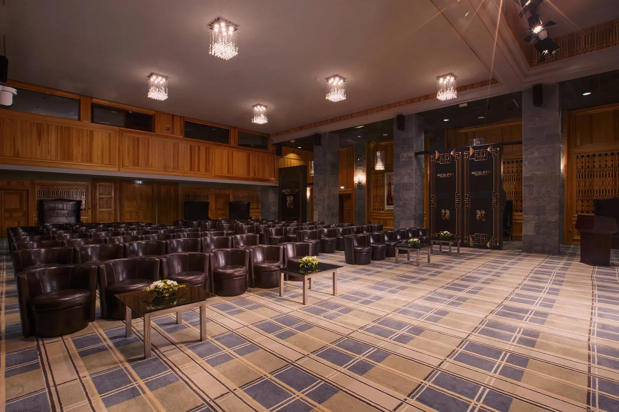 Salle de conférence avec des rangées de fauteuils en cuir marron, des tables basses avec des fleurs, des murs en bois et des lustres au plafond