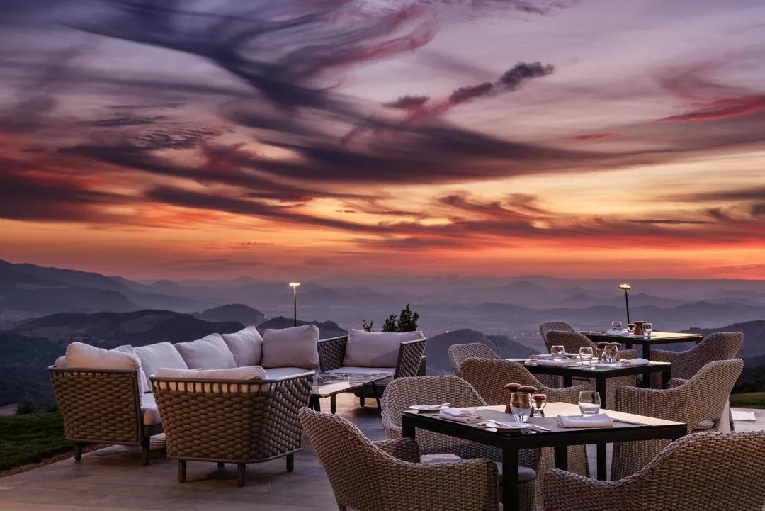 Terrasse avec des tables et des chaises en osier, des coussins blancs et des verres, face à un coucher de soleil sur des montagnes