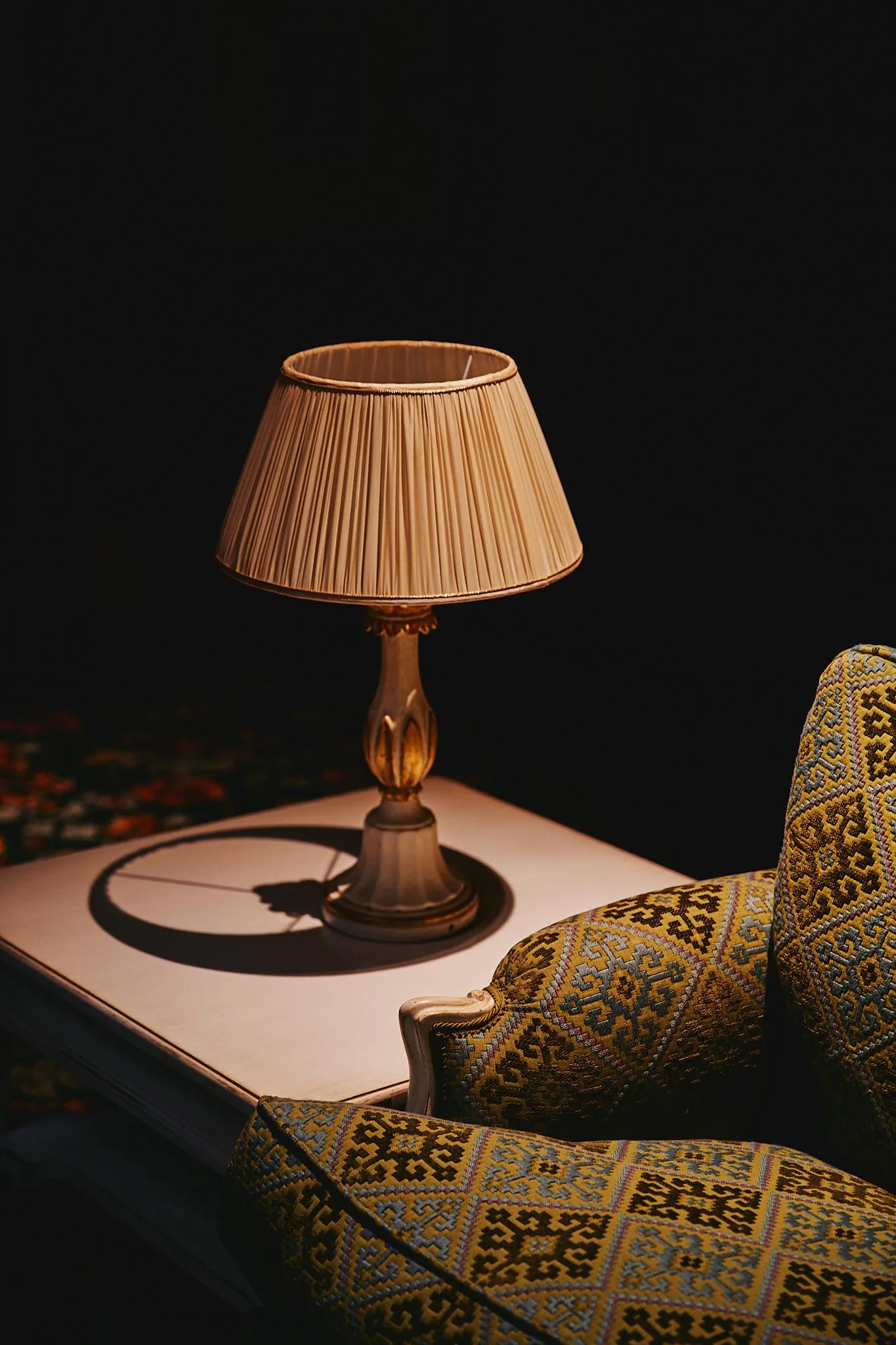 Une lampe à abat-jour beige éclairée est posée sur une table à côté d'un fauteuil aux motifs jaunes et bleus
