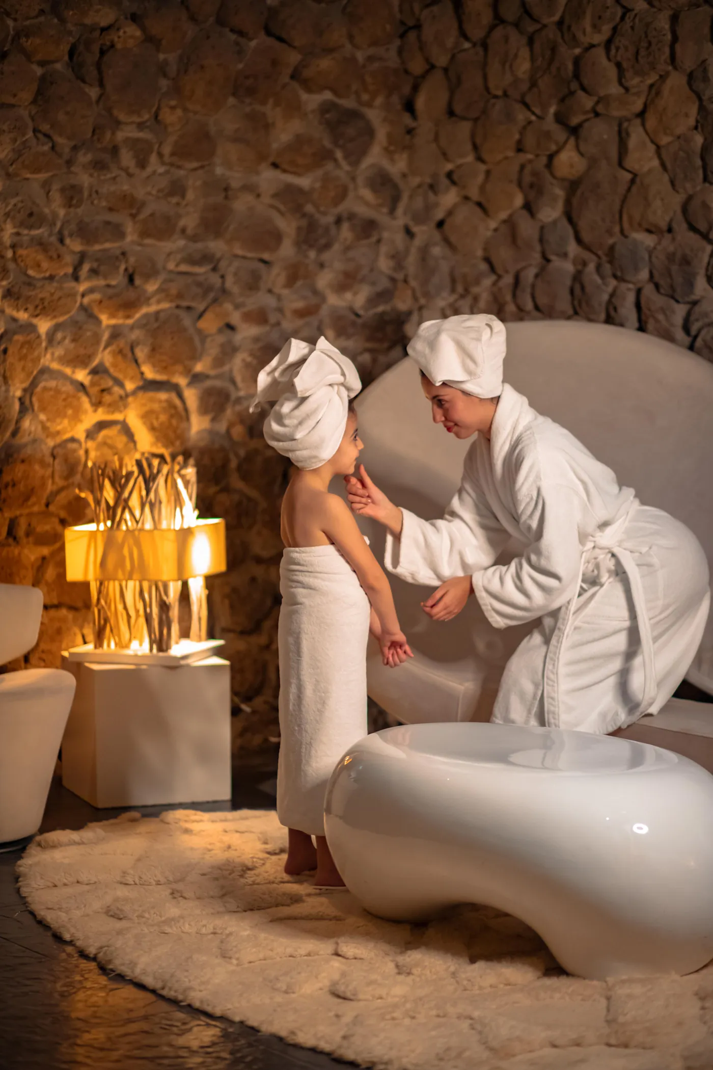 Une personne enveloppée dans une serviette et coiffée d'une serviette en turban est guidée par une autre personne vêtue d'une robe blanche dans un espace spa aux murs en pierre