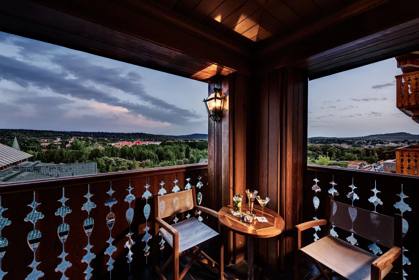Balcon en bois avec table, chaises et verres, offrant une vue panoramique sur une ville et des collines sous un ciel nuageux