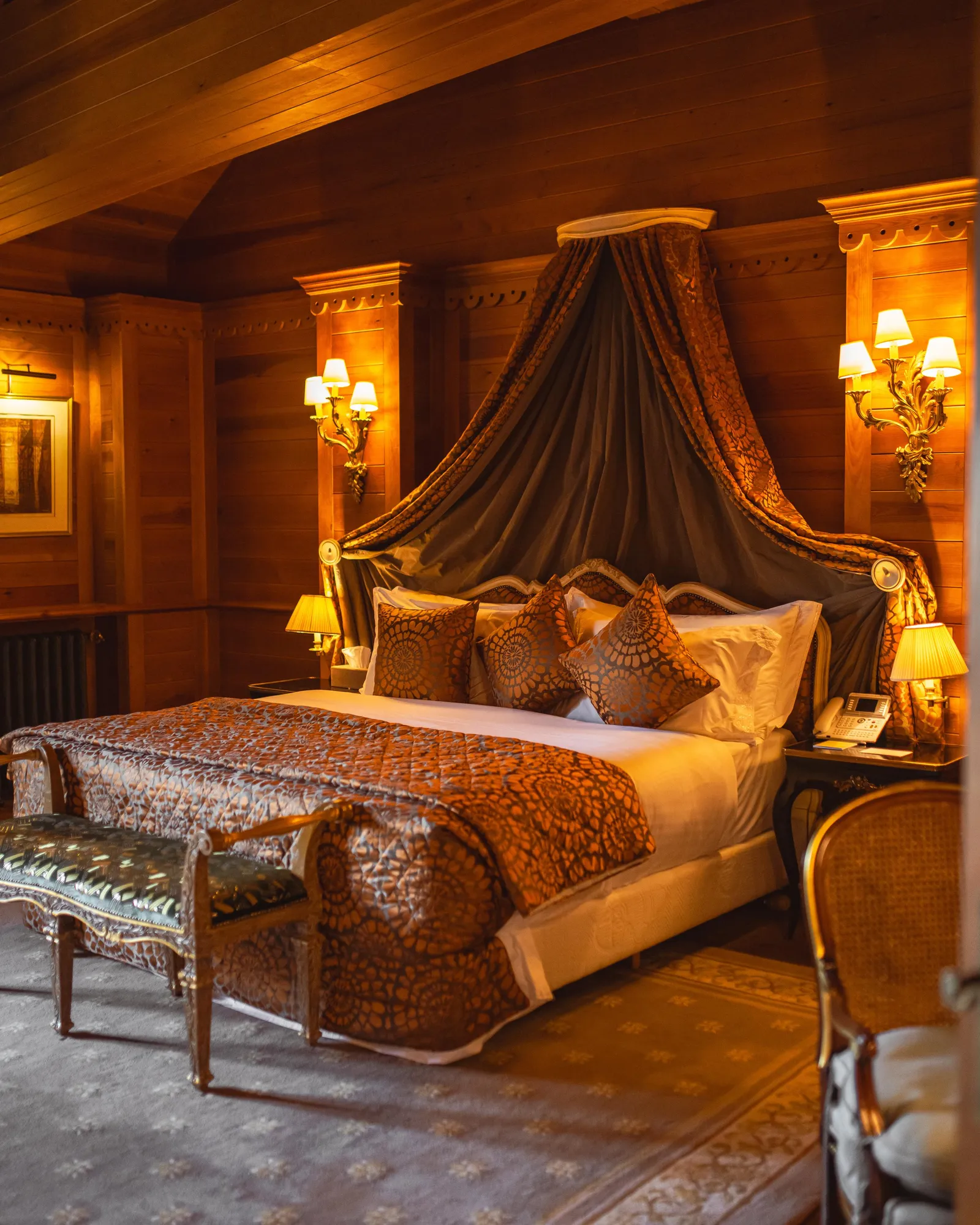 Chambre luxueuse avec un grand lit à baldaquin orné de rideaux et de coussins, encadrée par des boiseries et éclairée par des lampes murales et de chevet