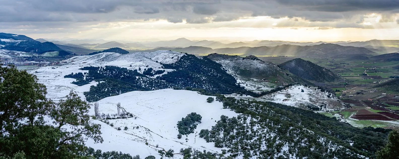 Paysage montagneux enneigé avec des collines, des champs et des forêts sous un ciel nuageux et ensoleillé