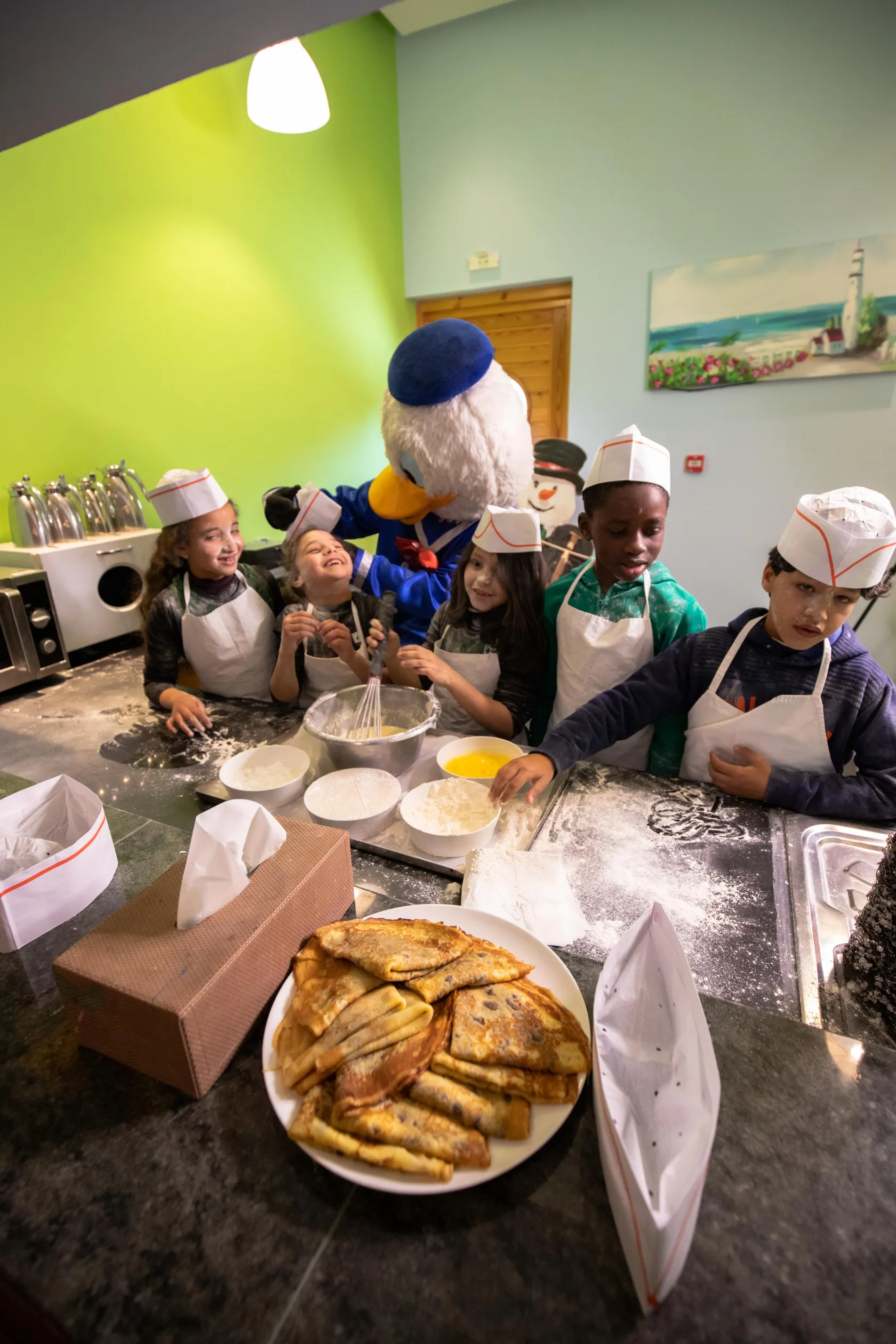 Un groupe d'enfants et un personnage mascottes cuisinent ensemble dans une cuisine, entourés d'ingrédients et d'ustensiles de pâtisserie