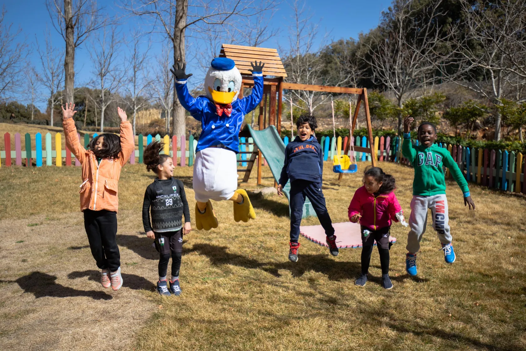 Un personnage costumé en canard saute avec cinq enfants dans un parc avec des jeux en bois et une clôture colorée en arrière-plan