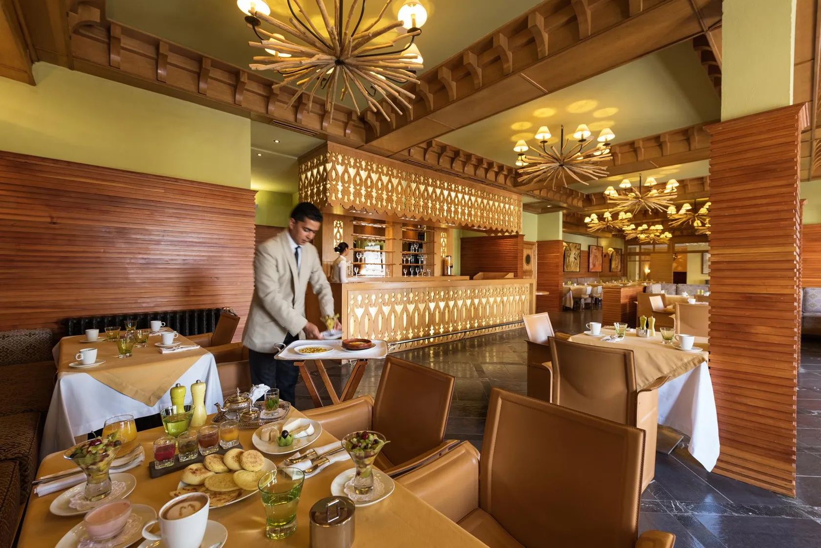 Un serveur en veste blanche porte un plateau avec des plats dans un restaurant élégant aux murs en bois et aux lustres décoratifs