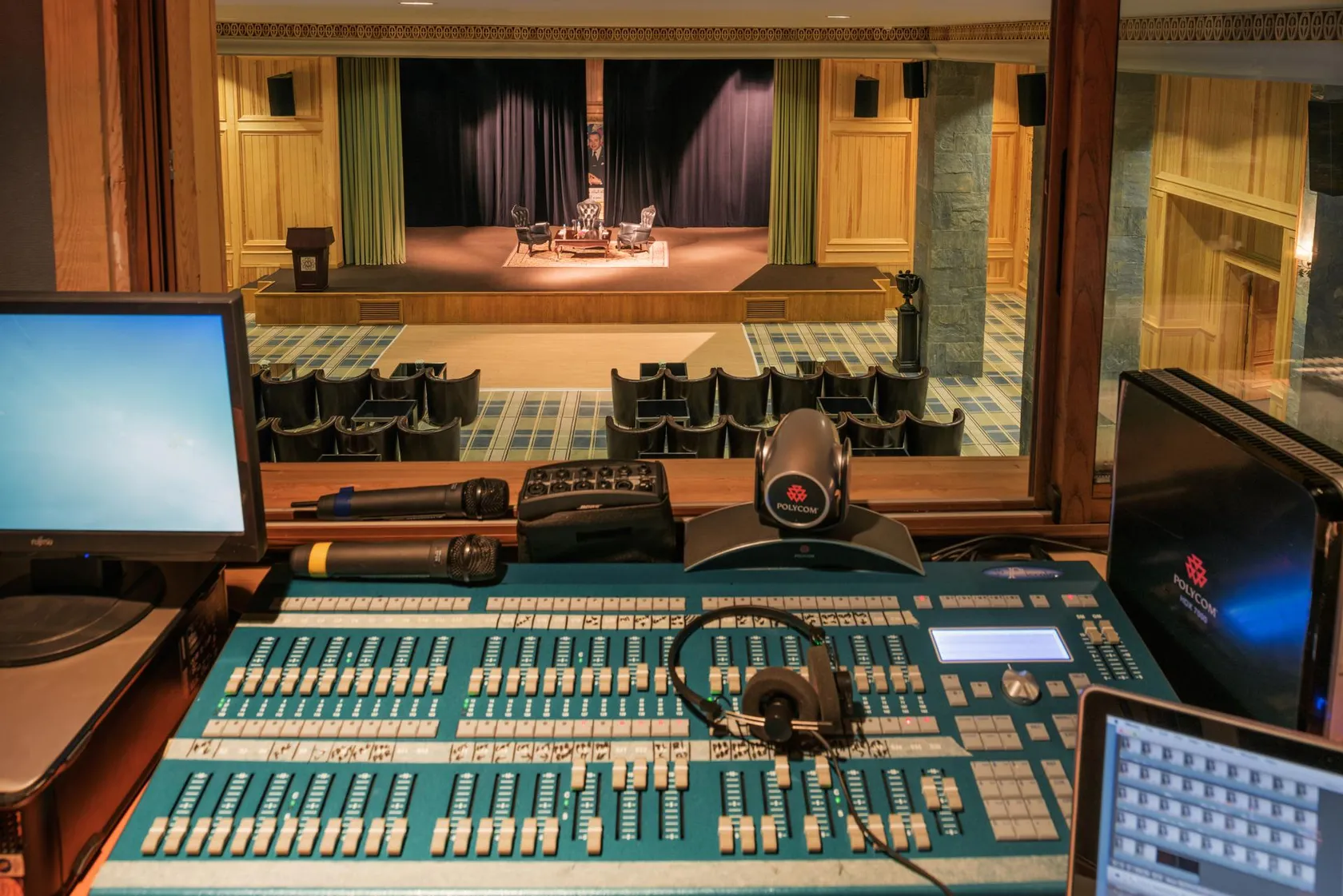 Table de mixage audio avec écrans et microphones devant une scène de théâtre avec des chaises et un rideau fermé