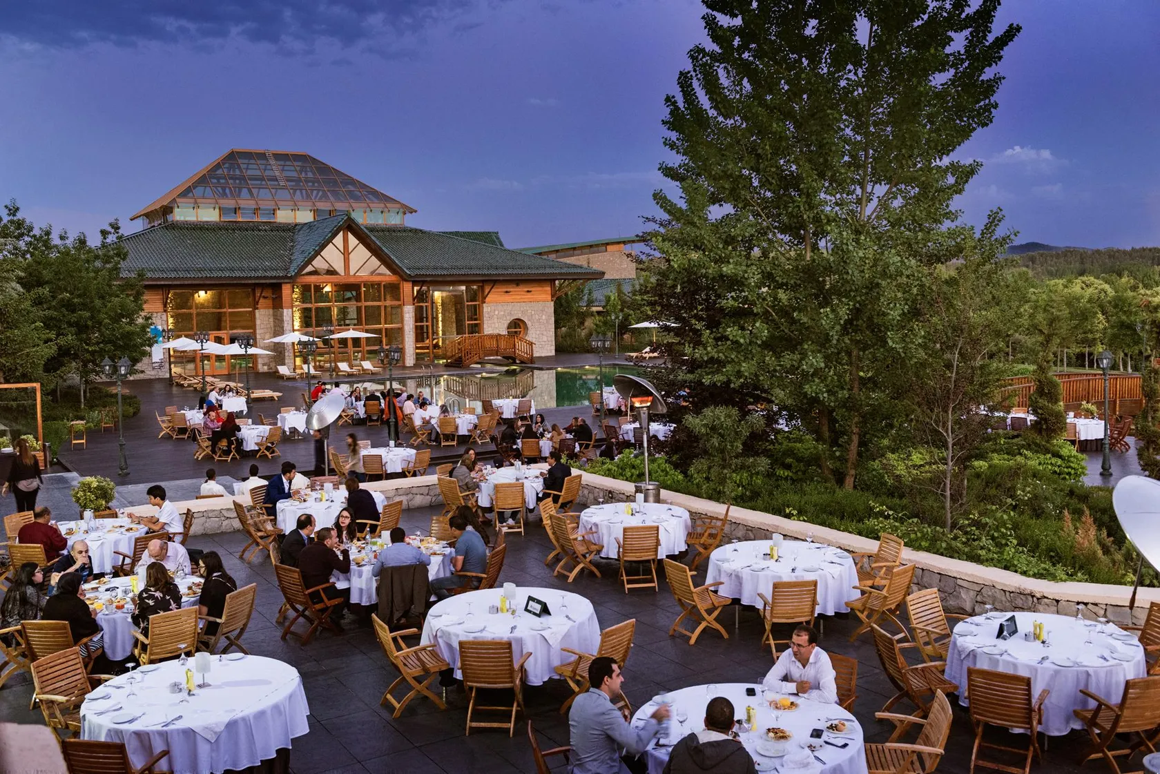 Une terrasse de restaurant en extérieur avec de nombreuses tables et chaises, des clients dînant et un bâtiment en bois et pierre en arrière-plan