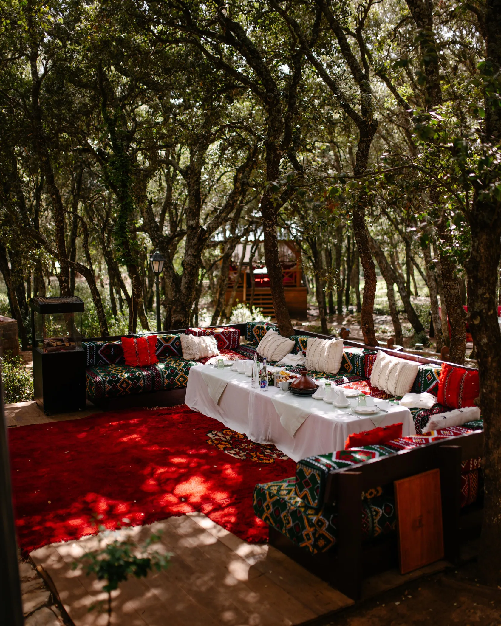 Un espace extérieur aménagé avec des coussins colorés, une table recouverte d'une nappe blanche et des arbres autour