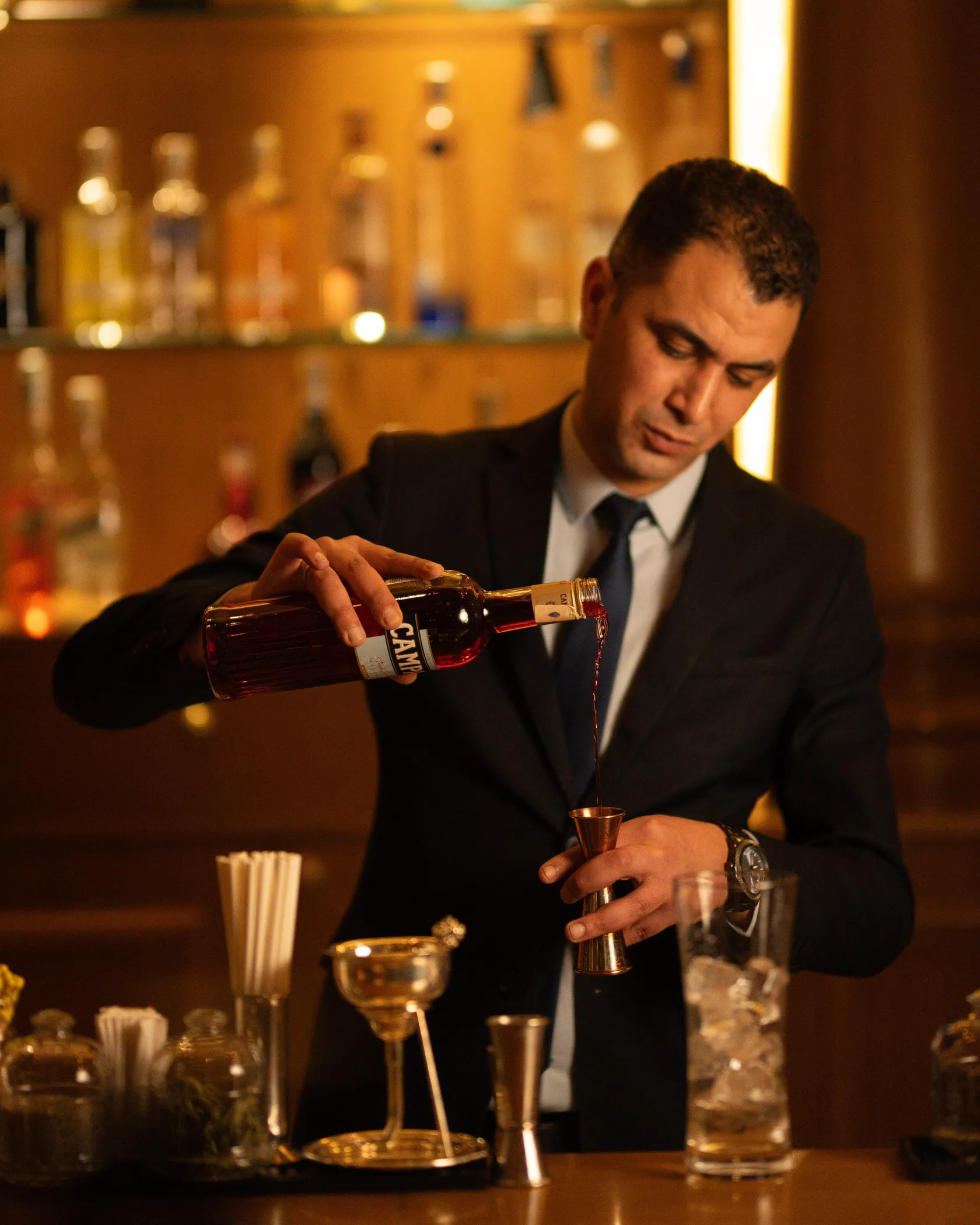 Un homme en costume verse un liquide d'une bouteille dans un petit verre métallique derrière un bar