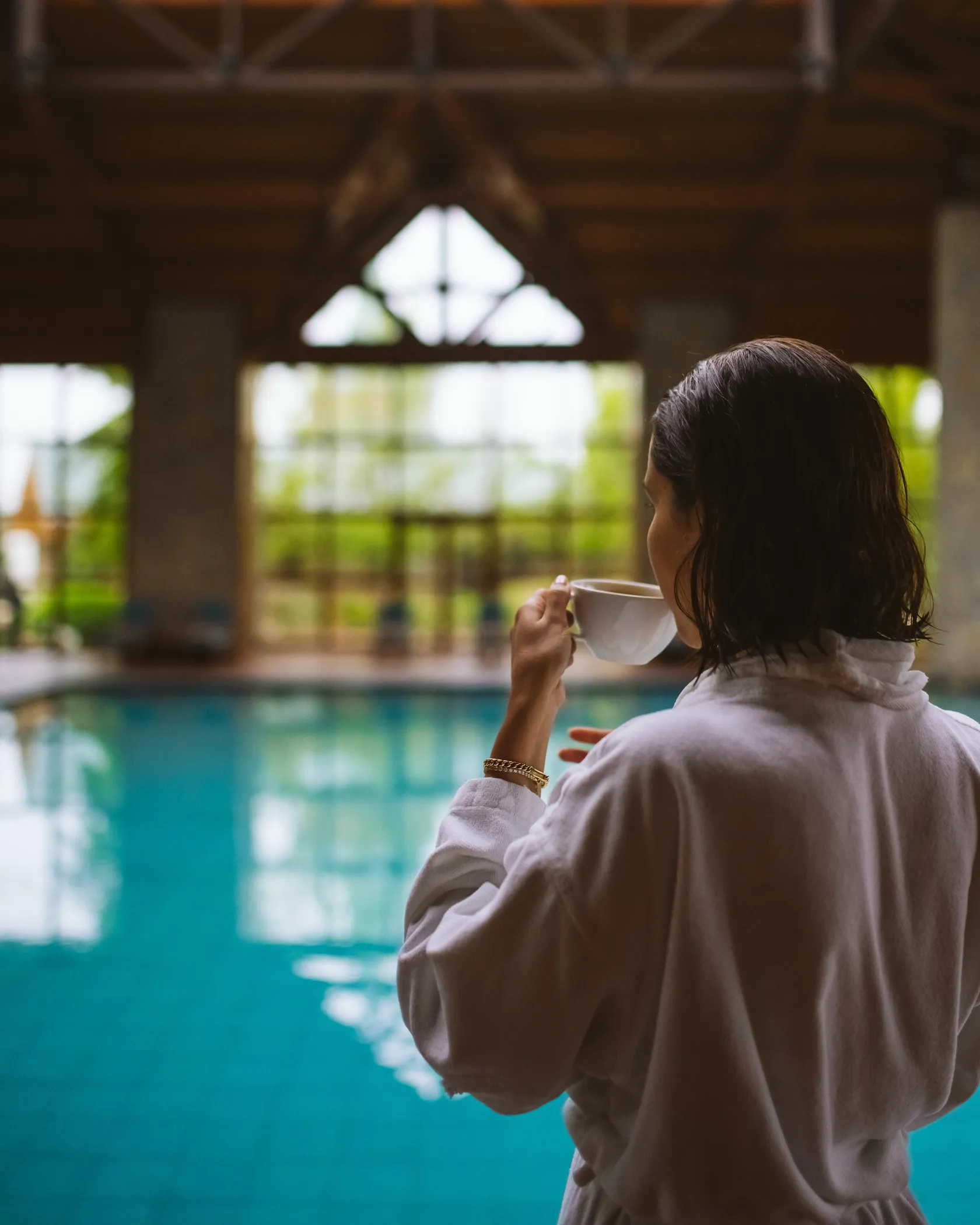 Une personne en robe de chambre tient une tasse près d'une piscine intérieure sous une structure en bois