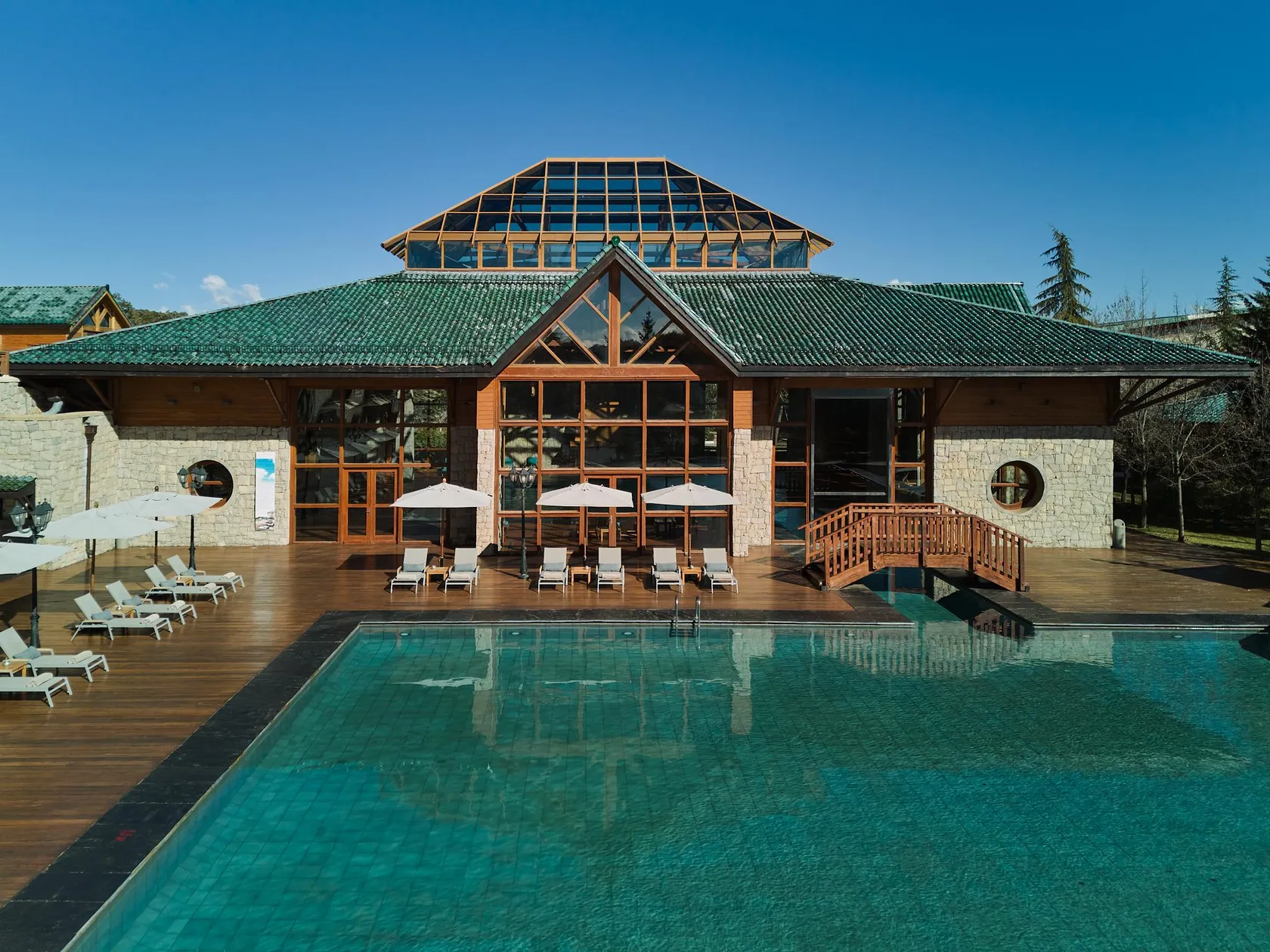 Un bâtiment en pierre et bois avec un toit vert et une extension vitrée, devant une piscine extérieure entourée de chaises longues et de parasols
