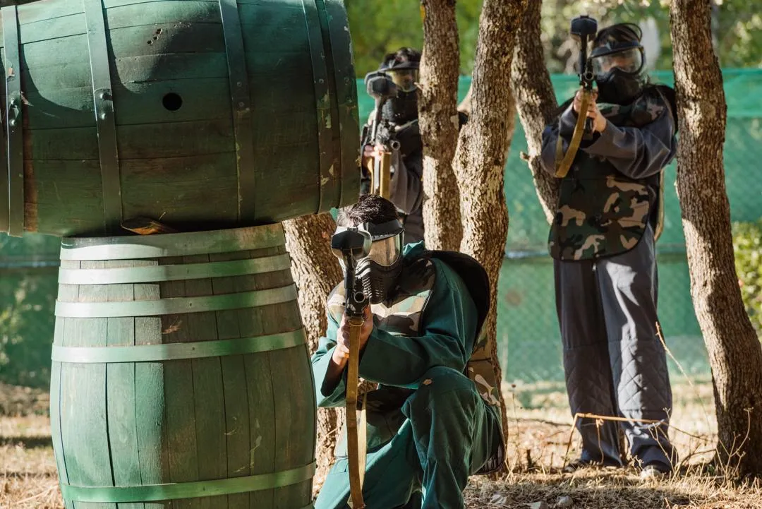 Trois personnes en tenue de camouflage et masques de protection se cachent derrière des barils en bois et des arbres, tenant des armes factices