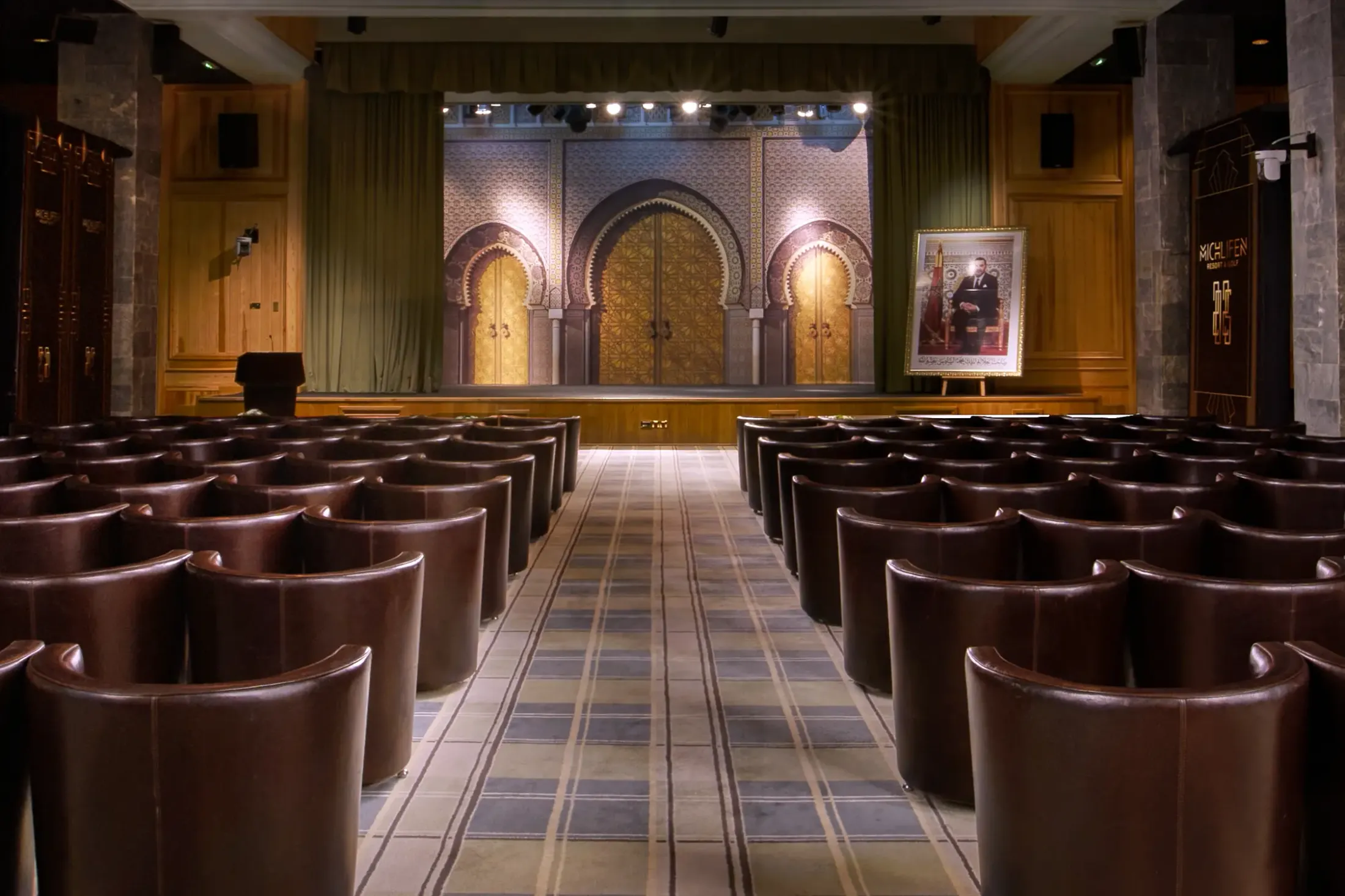 Salle de conférence avec des sièges en cuir disposés en rangées face à une scène ornée de motifs architecturaux et éclairée, avec un portrait sur un chevalet à droite