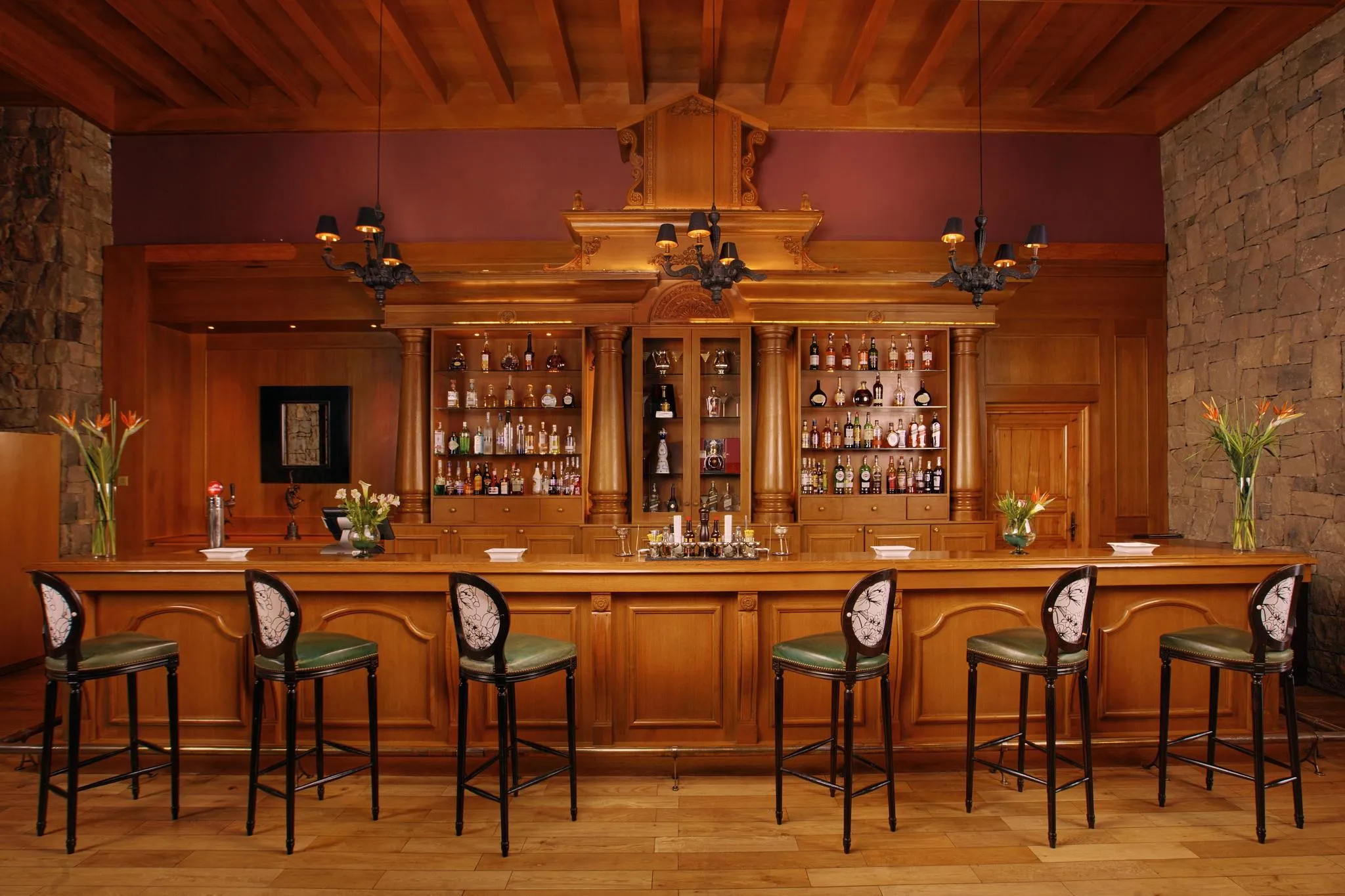 Un bar en bois avec des étagères remplies de bouteilles, des tabourets verts et des fleurs sur le comptoir