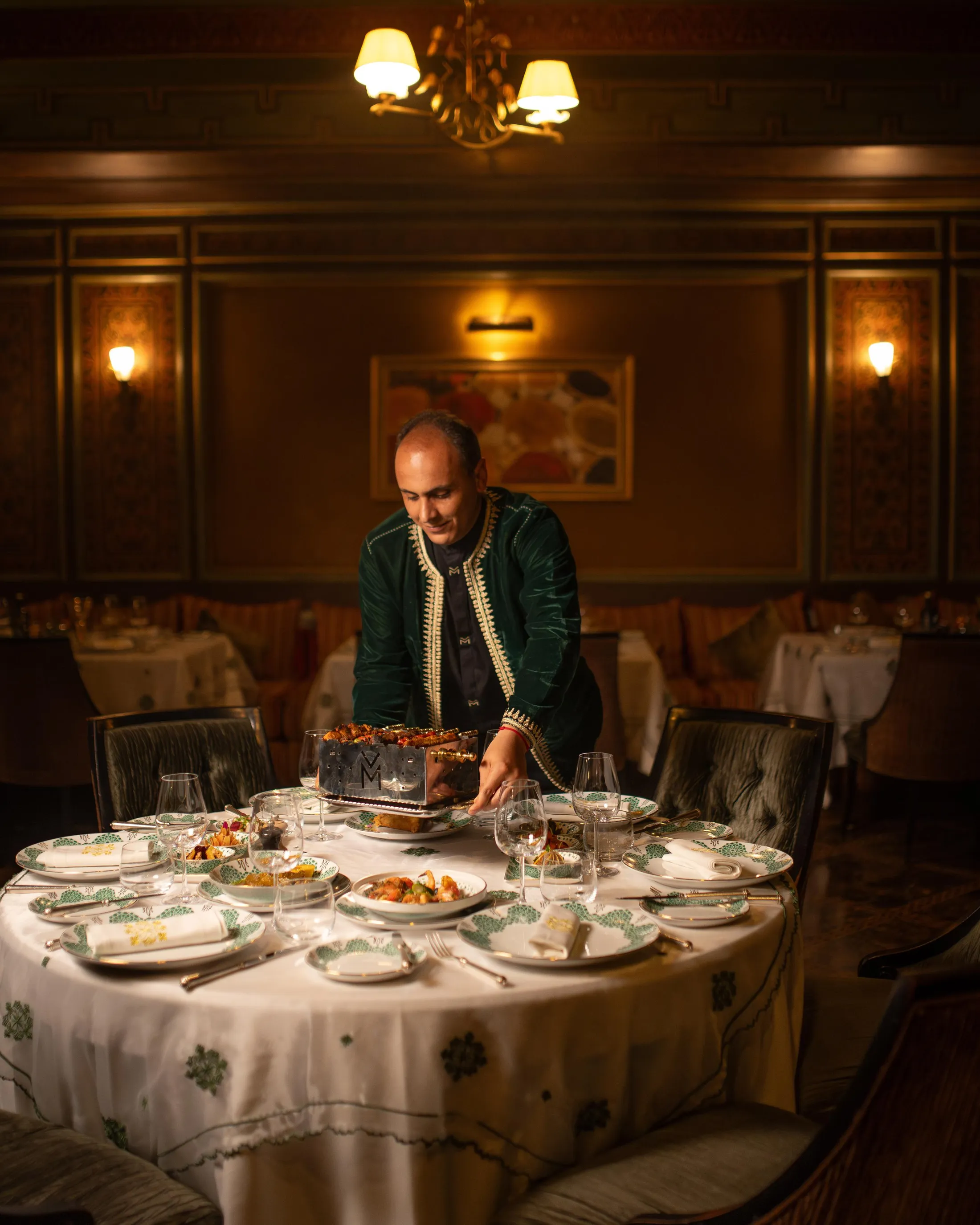 Un homme en veste verte sert un plat à un dîner formel dans une salle à manger élégante avec des chandeliers et des murs décorés