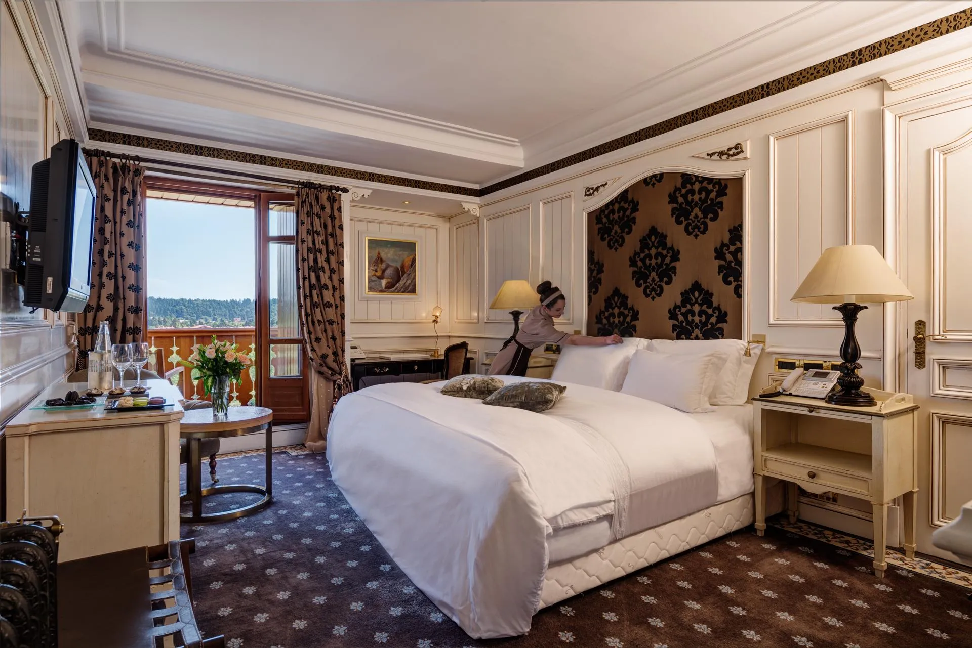Chambre d'hôtel luxueuse avec un lit double, une fenêtre offrant une vue sur un paysage, des tables de chevet avec lampes et un téléviseur mural