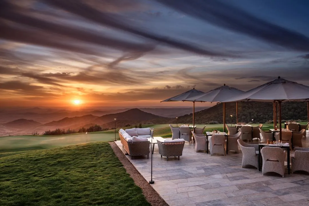Terrasse avec des chaises et des tables sous des parasols, face à un coucher de soleil sur des montagnes et un terrain de golf