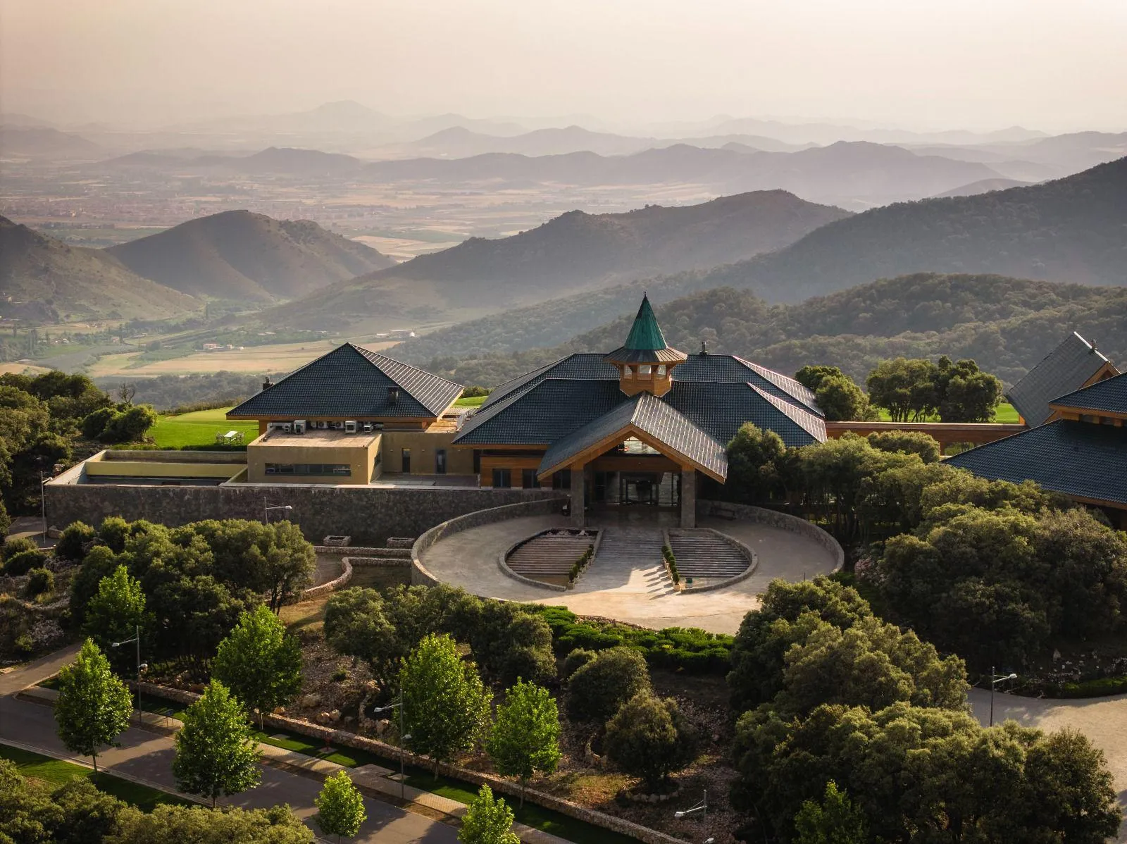 Un bâtiment avec des toits pentus et une tour centrale se trouve dans un paysage vallonné et verdoyant, entouré d'arbres et de jardins