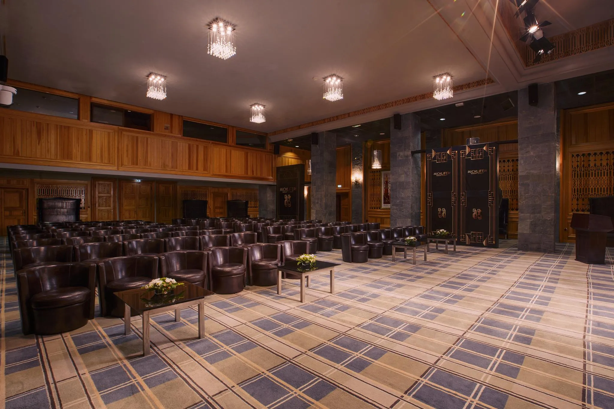 Salle de conférence avec des rangées de fauteuils en cuir marron, des tables basses avec des fleurs, des murs en bois et des lustres au plafond