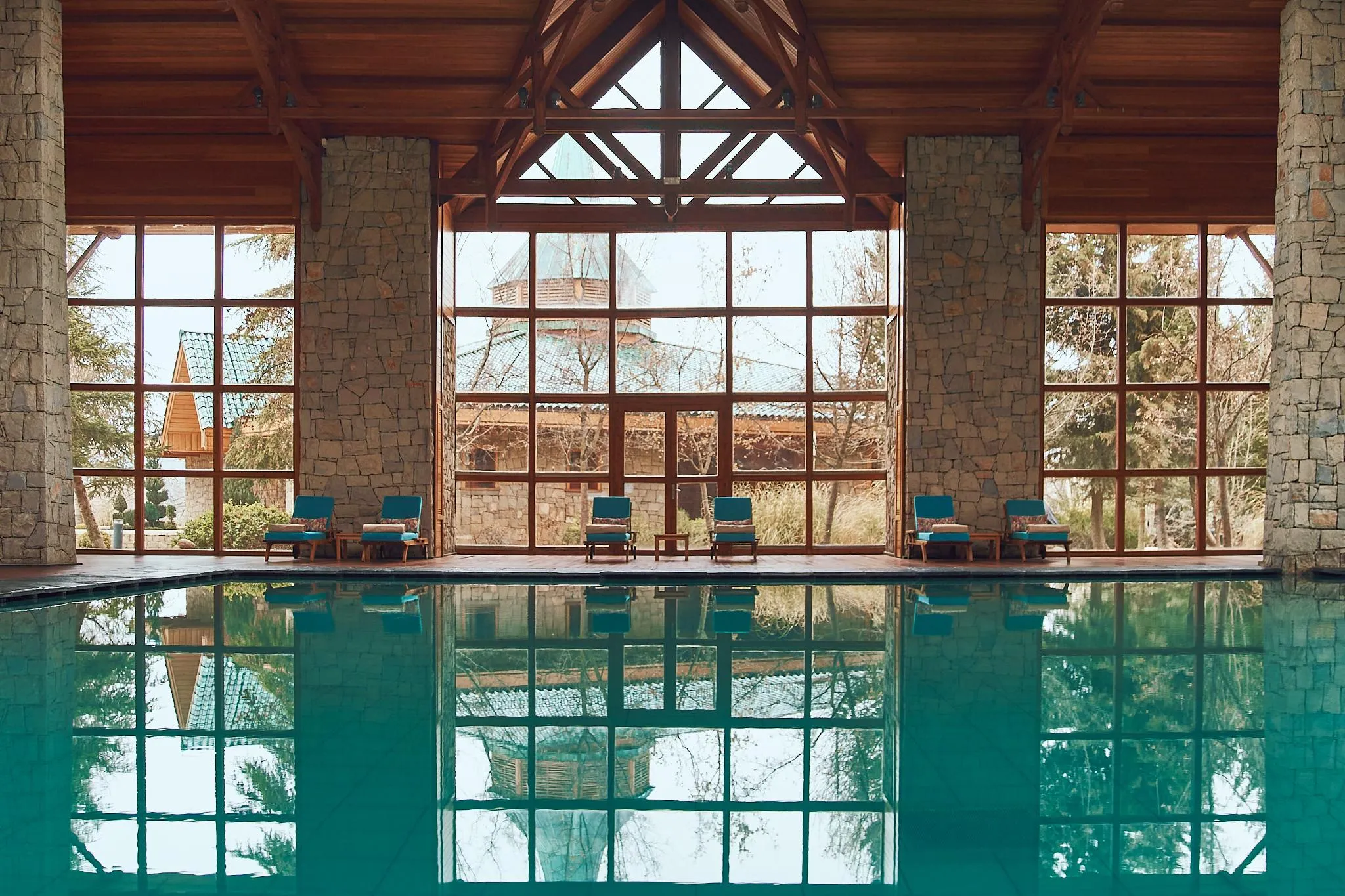 Piscine intérieure avec vue sur des chaises longues et des structures en pierre et bois à travers de grandes fenêtres