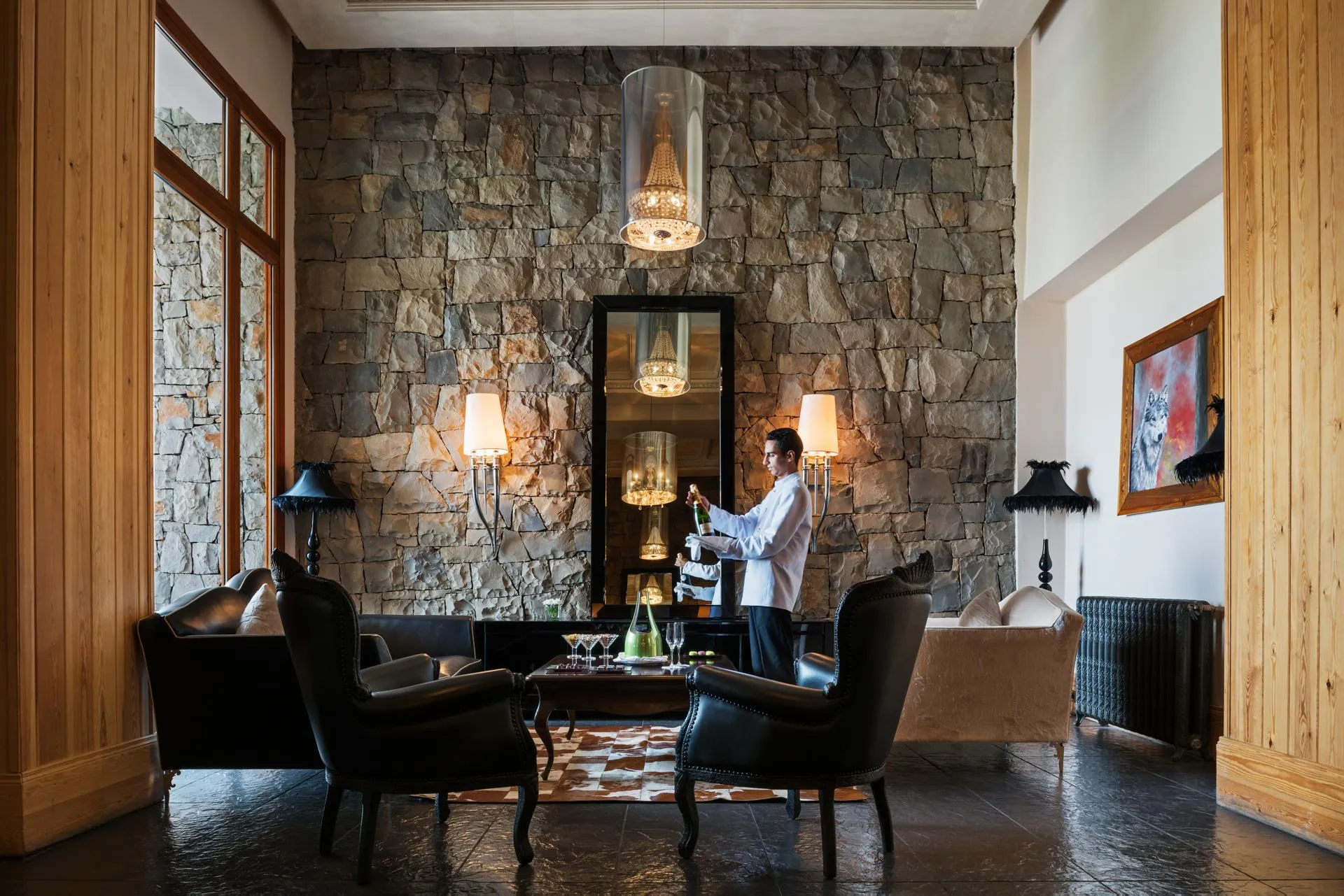 Un serveur en veste blanche prépare des boissons derrière un bar en pierre avec des chandeliers, entouré de fauteuils et de lampes dans un salon élégant