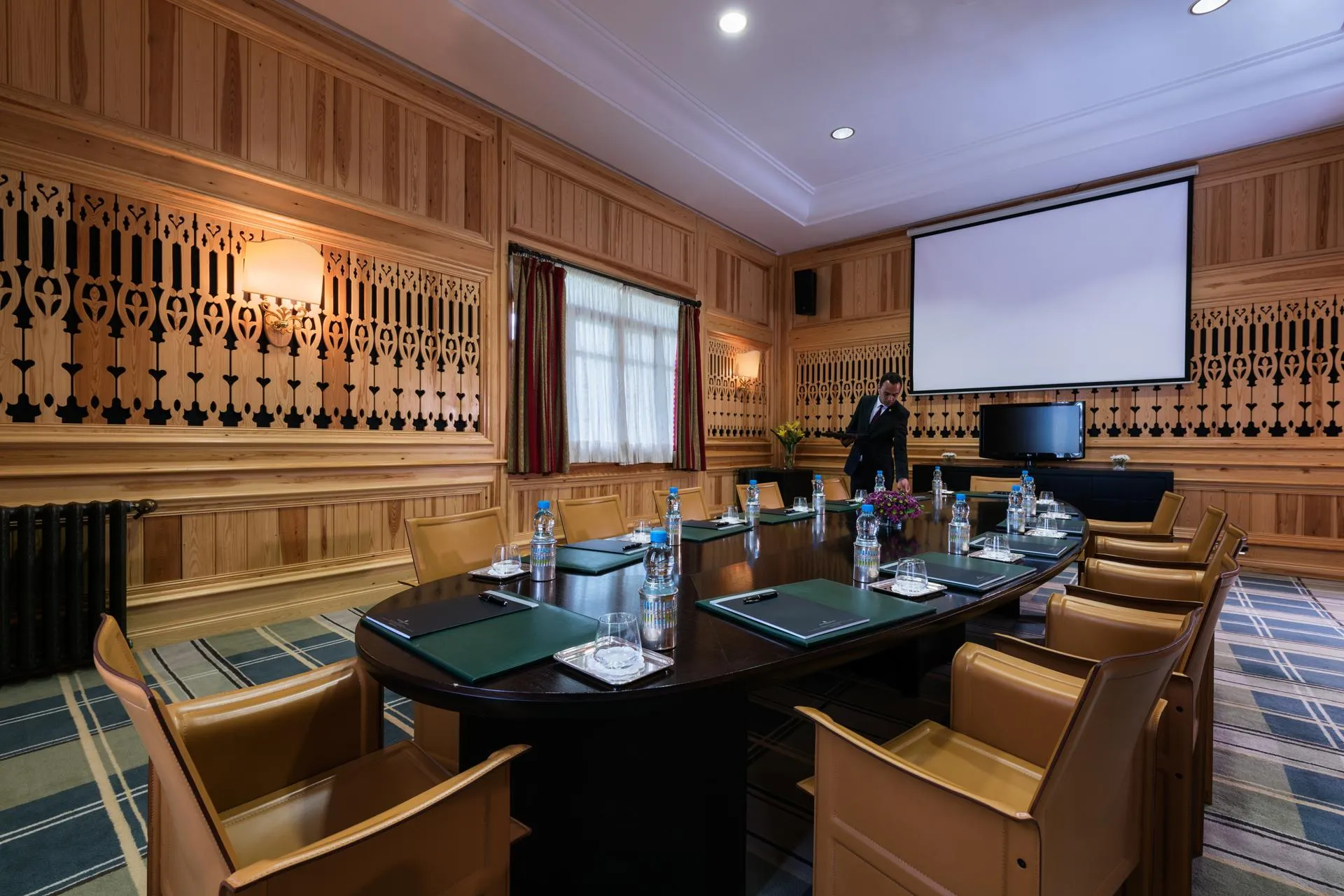 Salle de réunion en bois avec une table ovale, des chaises en cuir, un projecteur et un homme debout près d'un écran