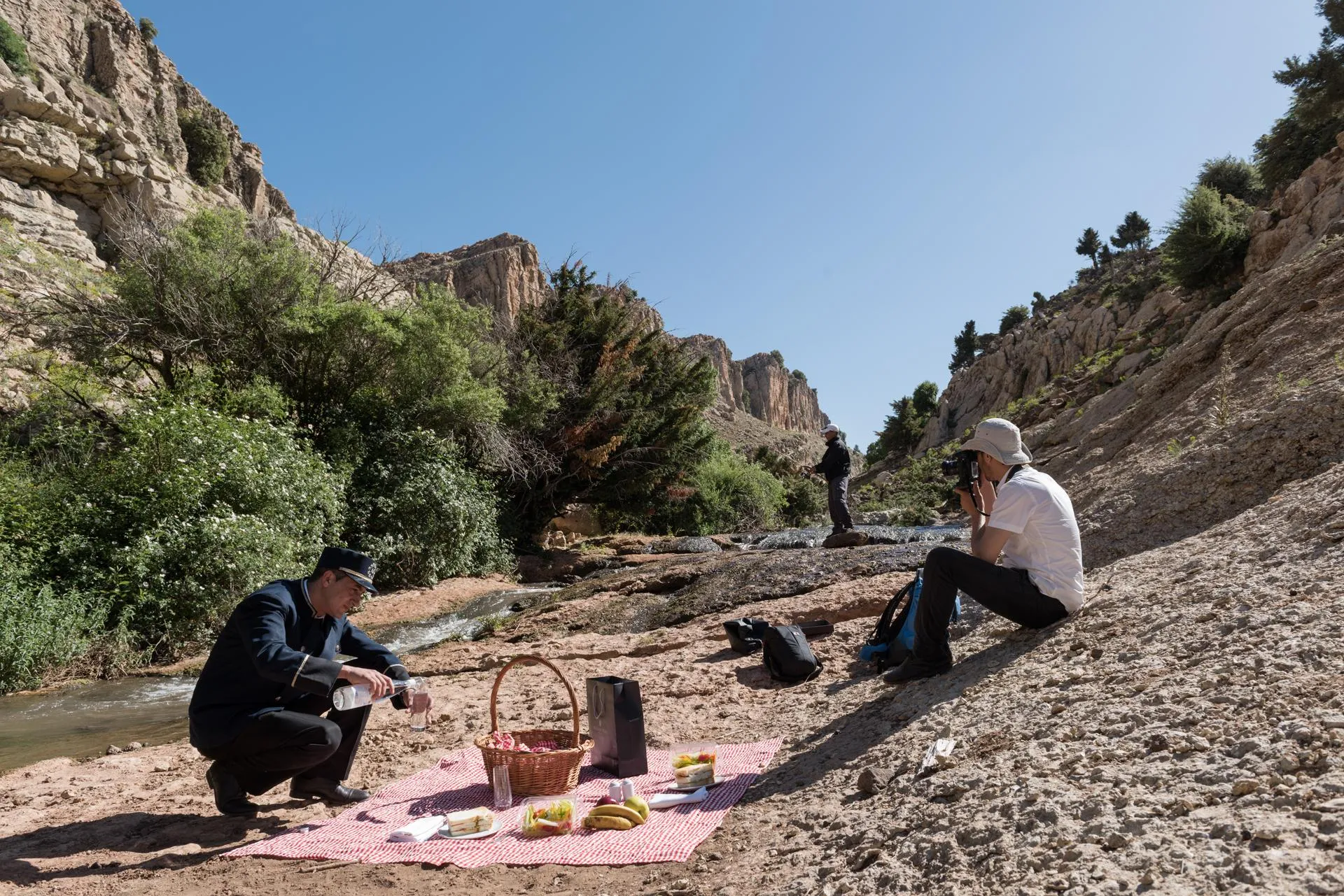 Deux personnes pique-niquent près d'un ruisseau dans un canyon rocheux, avec des fruits, une bouteille et un panier sur une couverture