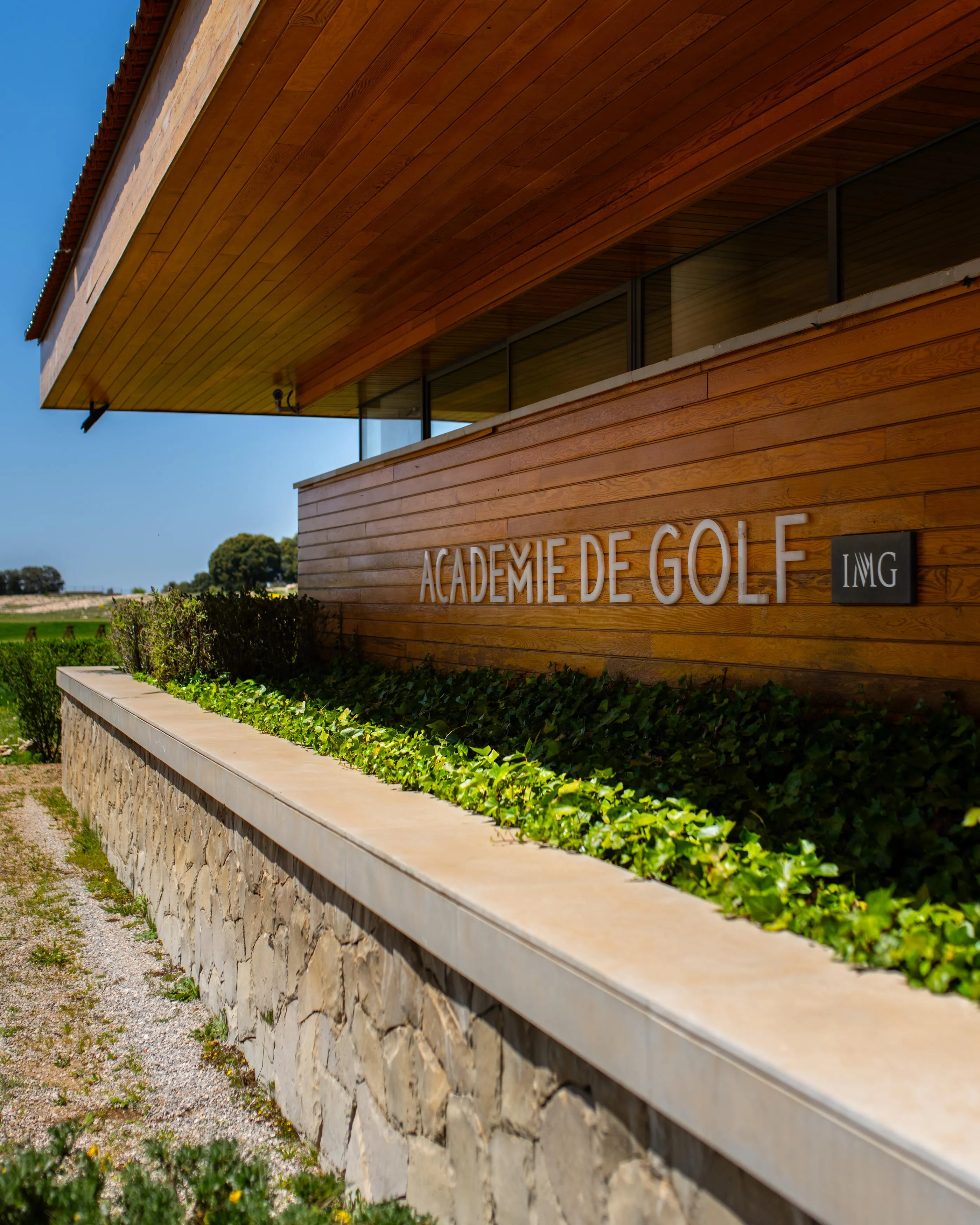Wooden building with the inscription "IMG Golf Academy" and a stone wall with green plants