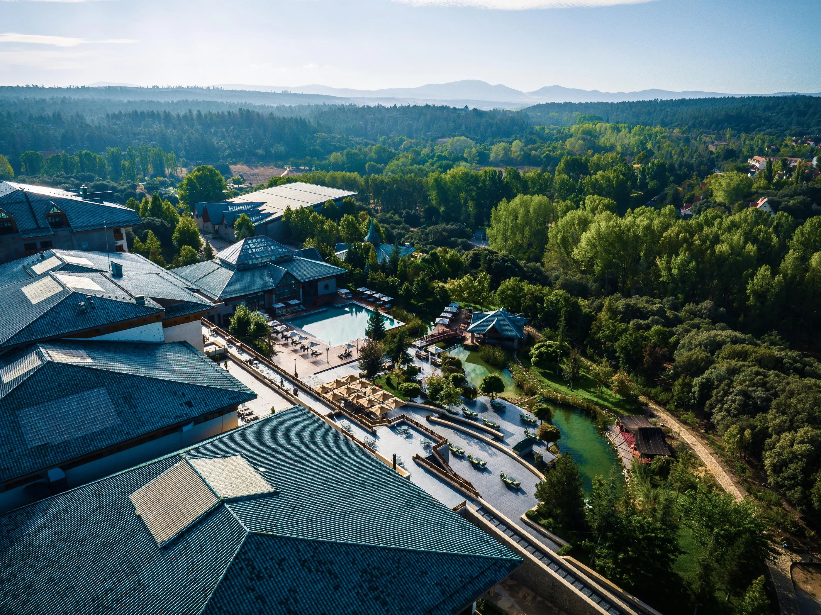 Vue aérienne d'un complexe de bâtiments traditionnels avec toits en tuiles, entouré d'arbres et de plans d'eau, dans un paysage montagneux