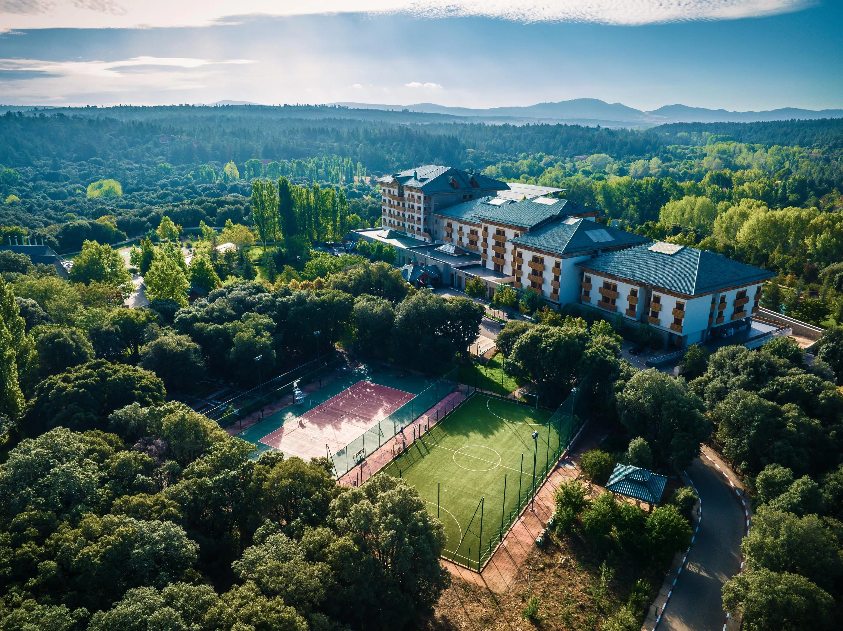 Vue aérienne d'un bâtiment entouré d'arbres avec des courts de tennis et un terrain de sport en contrebas, dans un paysage forestier et montagneux