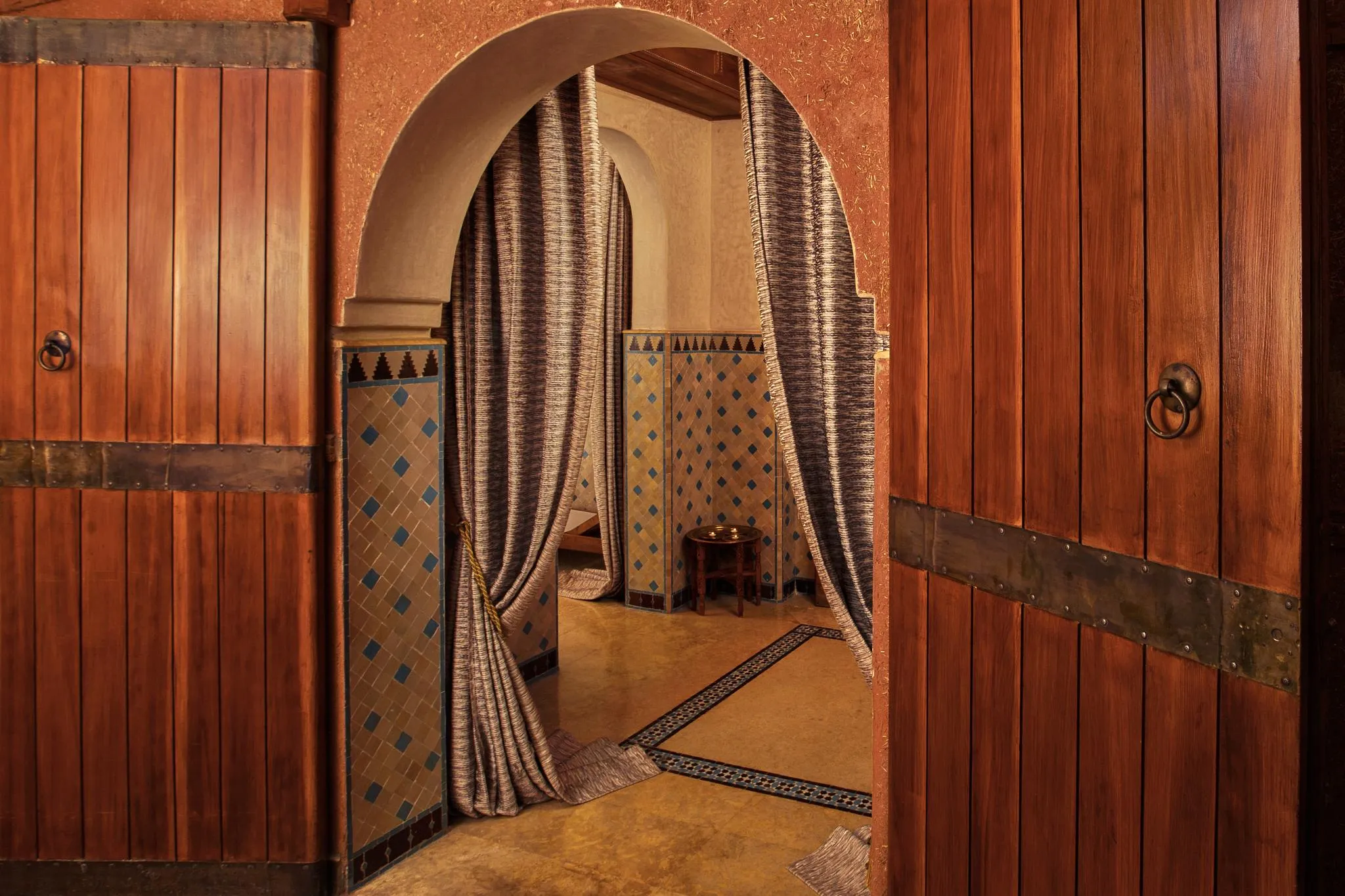 Deux portes en bois avec des anneaux en métal ouvrent sur une pièce aux murs carrelés et aux arches drapées de tissu