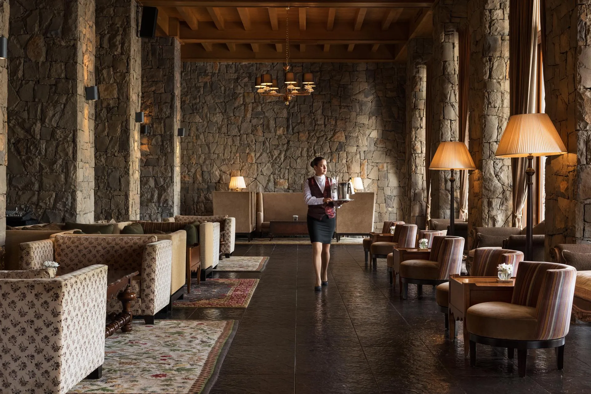 Une personne en uniforme de serveur traverse une salle avec des canapés, des fauteuils et des lampes, dans un cadre aux murs en pierre et aux poutres apparentes