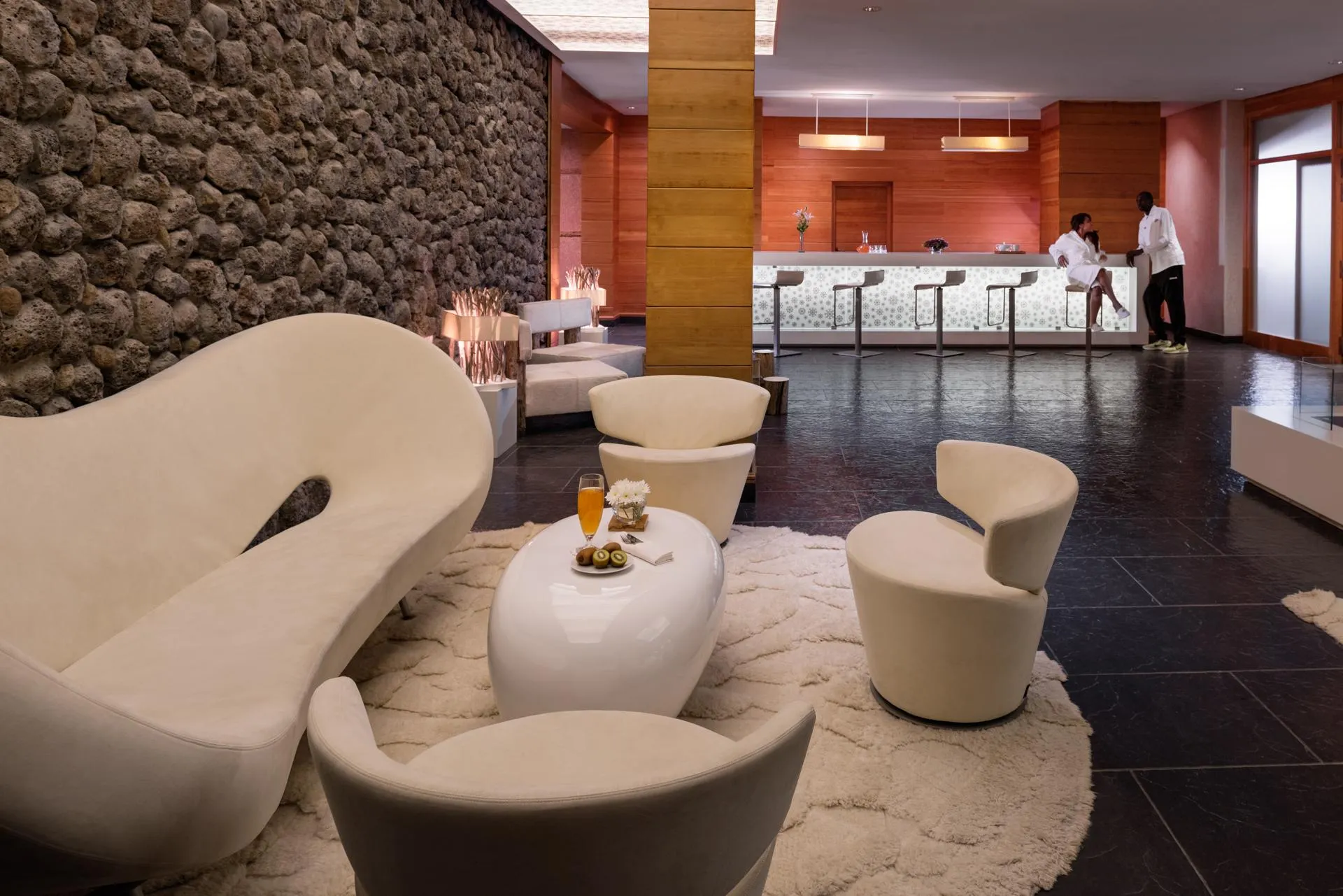 Un salon d'hôtel avec des sièges modernes, une table basse avec des boissons et des fruits, et un bar avec des tabourets en arrière-plan