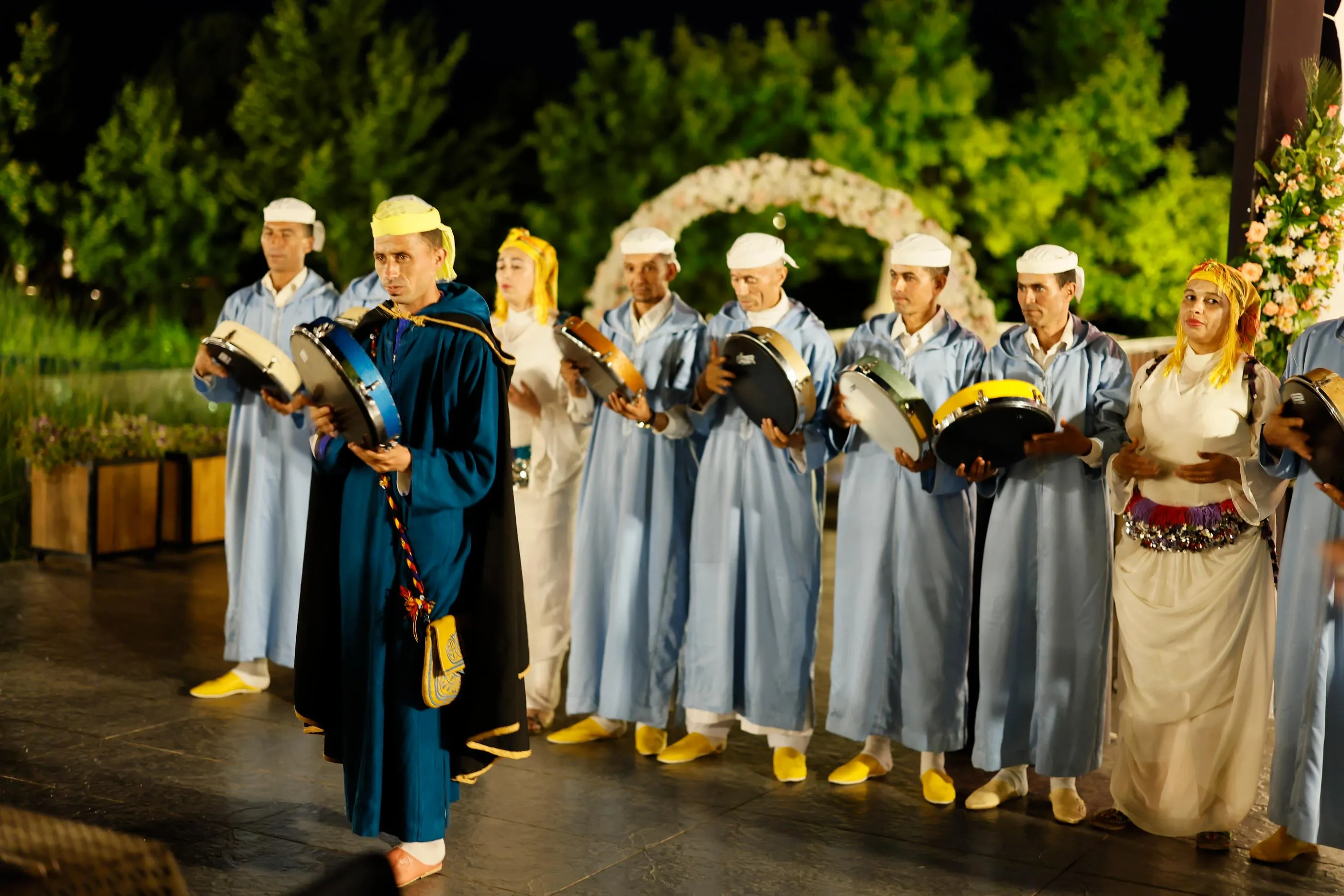 Un groupe de personnes en costumes traditionnels joue de petits tambours sur une scène extérieure décorée de fleurs