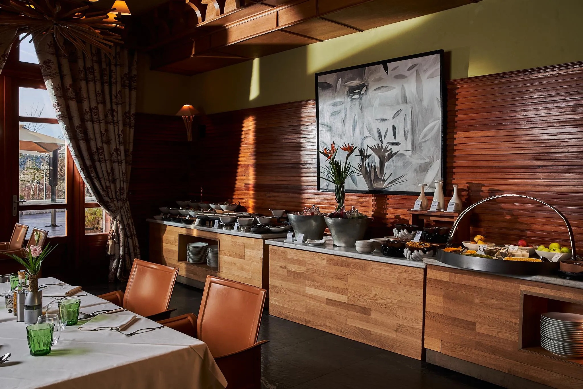 Un buffet de petit-déjeuner avec des fruits, des boissons et des plats chauds dans un restaurant aux murs en bois et aux grandes fenêtres