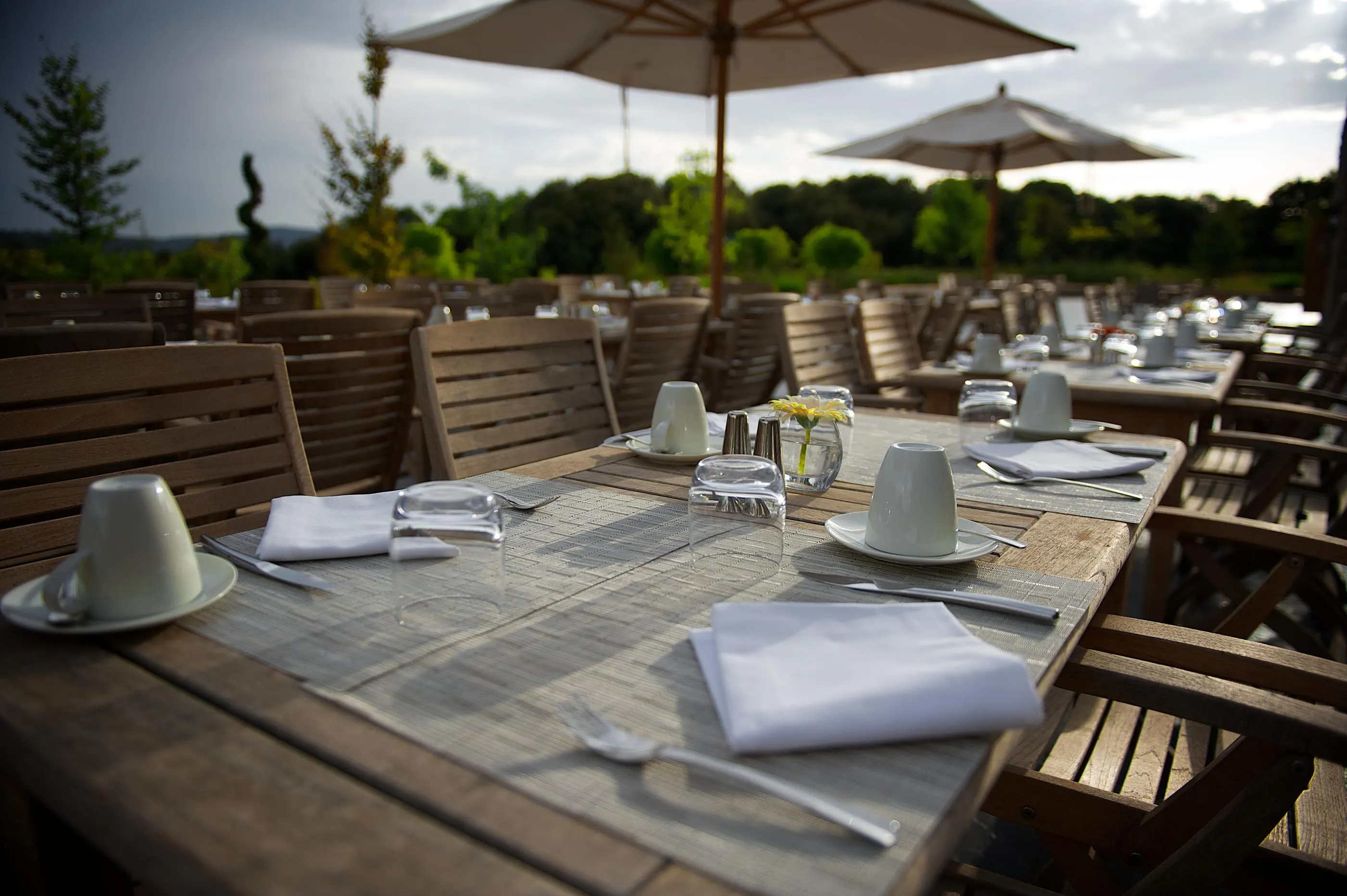 Table en terrasse extérieure avec vaisselle, verres et serviettes, entourée de chaises en bois et ombragée par des parasols