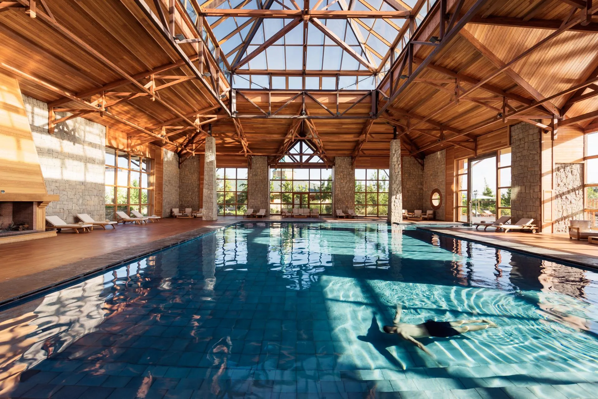 Piscine intérieure avec un toit en bois et des grandes fenêtres, entourée de chaises longues et de colonnes en pierre