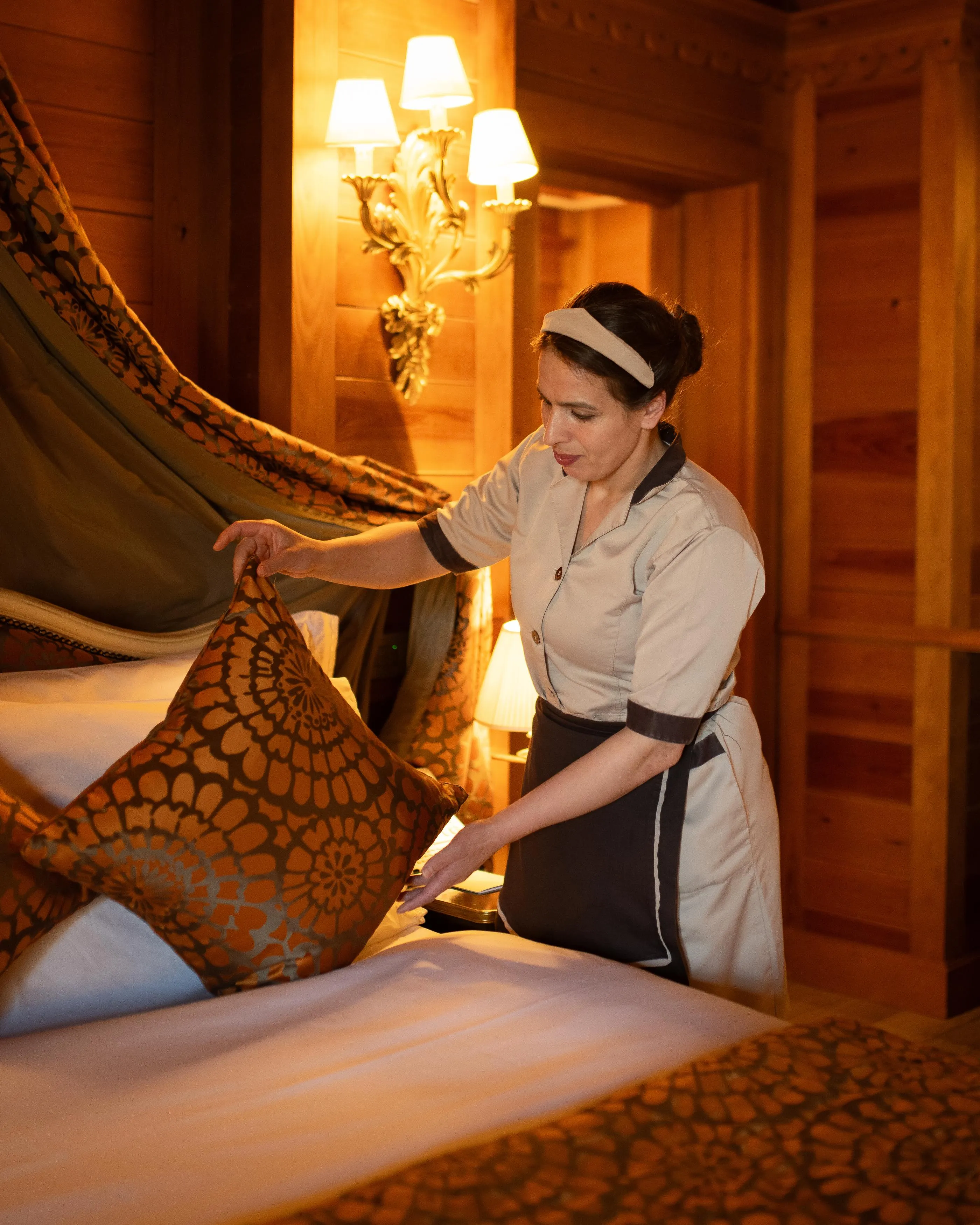 Une femme en uniforme d'employée de maison arrange des coussins sur un lit dans une chambre à la décoration ancienne